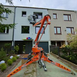 Auto-Klim Konrad Nowak - Montaż zewnętrznej jednostki klimatyzacji na elewacji domu z użyciem podnośnika koszowego firmy Boels, widok z dołu na pracownika w koszu na tle okna.