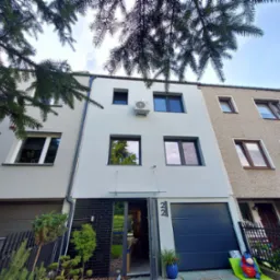 Elewacja nowoczesnego szeregowca z zamontowaną klimatyzacją na wysokości pierwszego piętra, widok z dołu przez gałęzie drzewa.