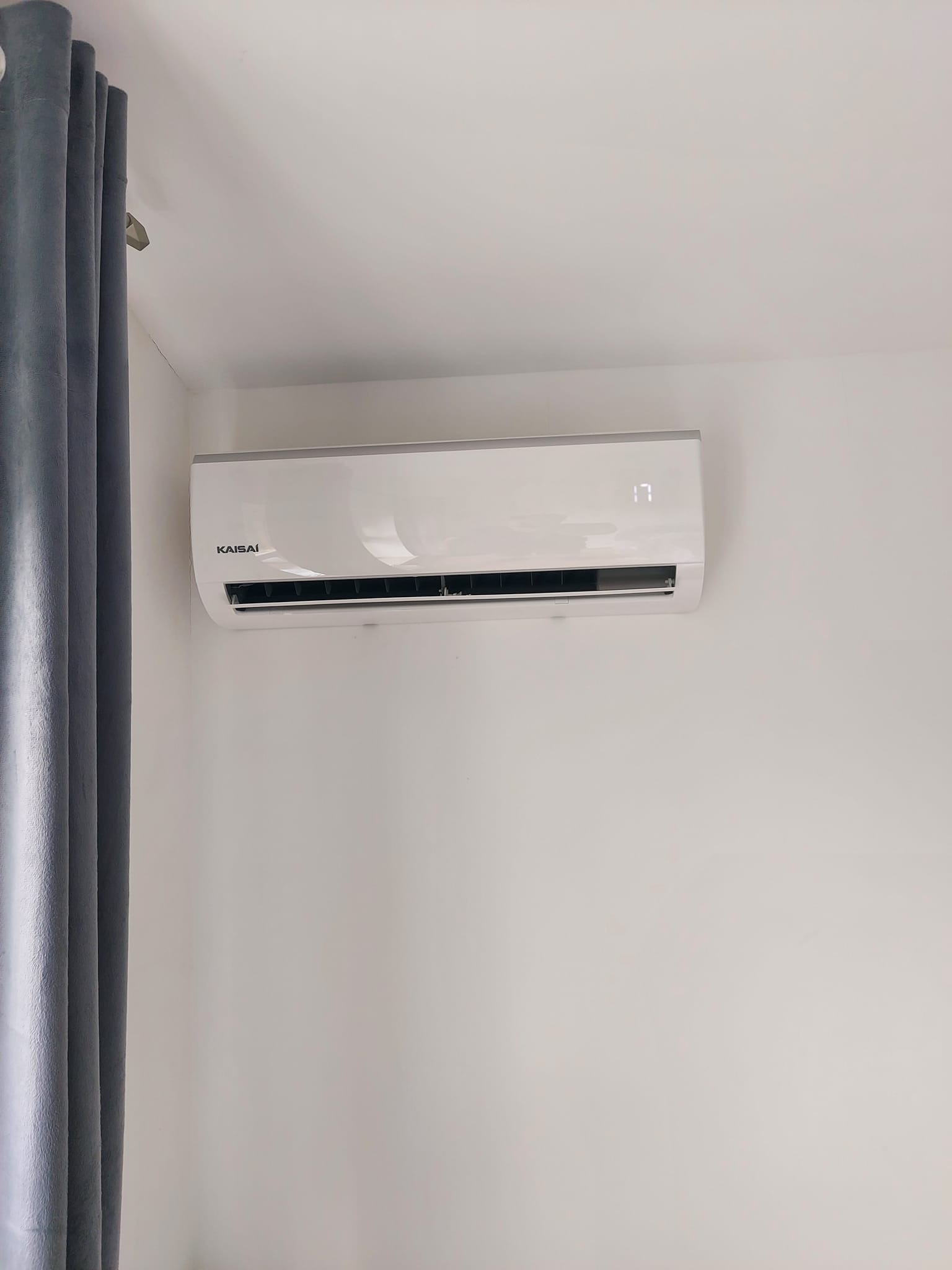 Biała jednostka klimatyzacji Kaisaí zamontowana na jasnej ścianie, z wyświetlaczem wskazującym temperaturę 17 stopni, obok zasłony w odcieniach szarości.