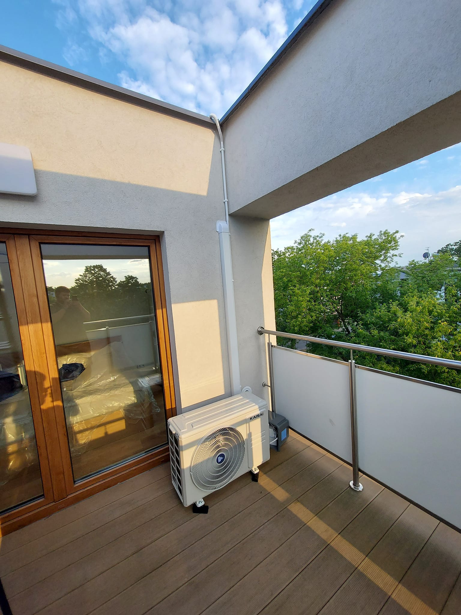 Jednostka zewnętrzna klimatyzacji Kaisai na balkonie z drewnianą podłogą, widok na zieleń, częściowo osłonięta folią okienna rama odbija fotografa.