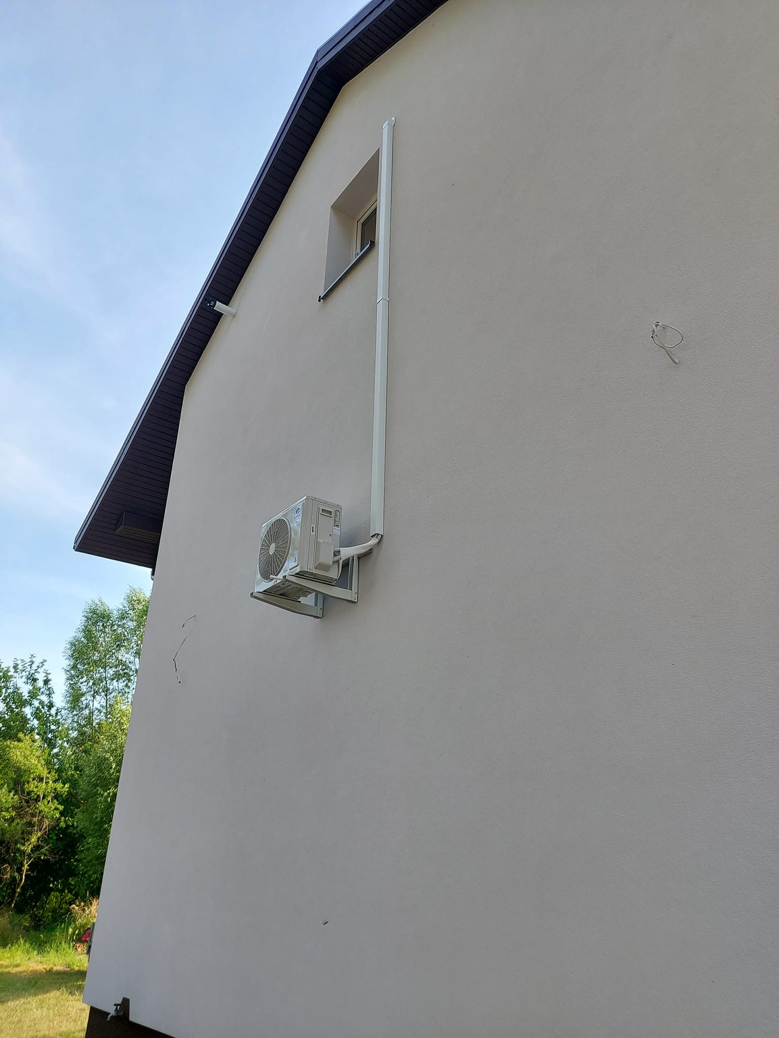 Zewnętrzna jednostka klimatyzacji zamontowana na ścianie domu jednorodzinnego, z widocznymi rurami odprowadzającymi skropliny, ujęcie z dołu.