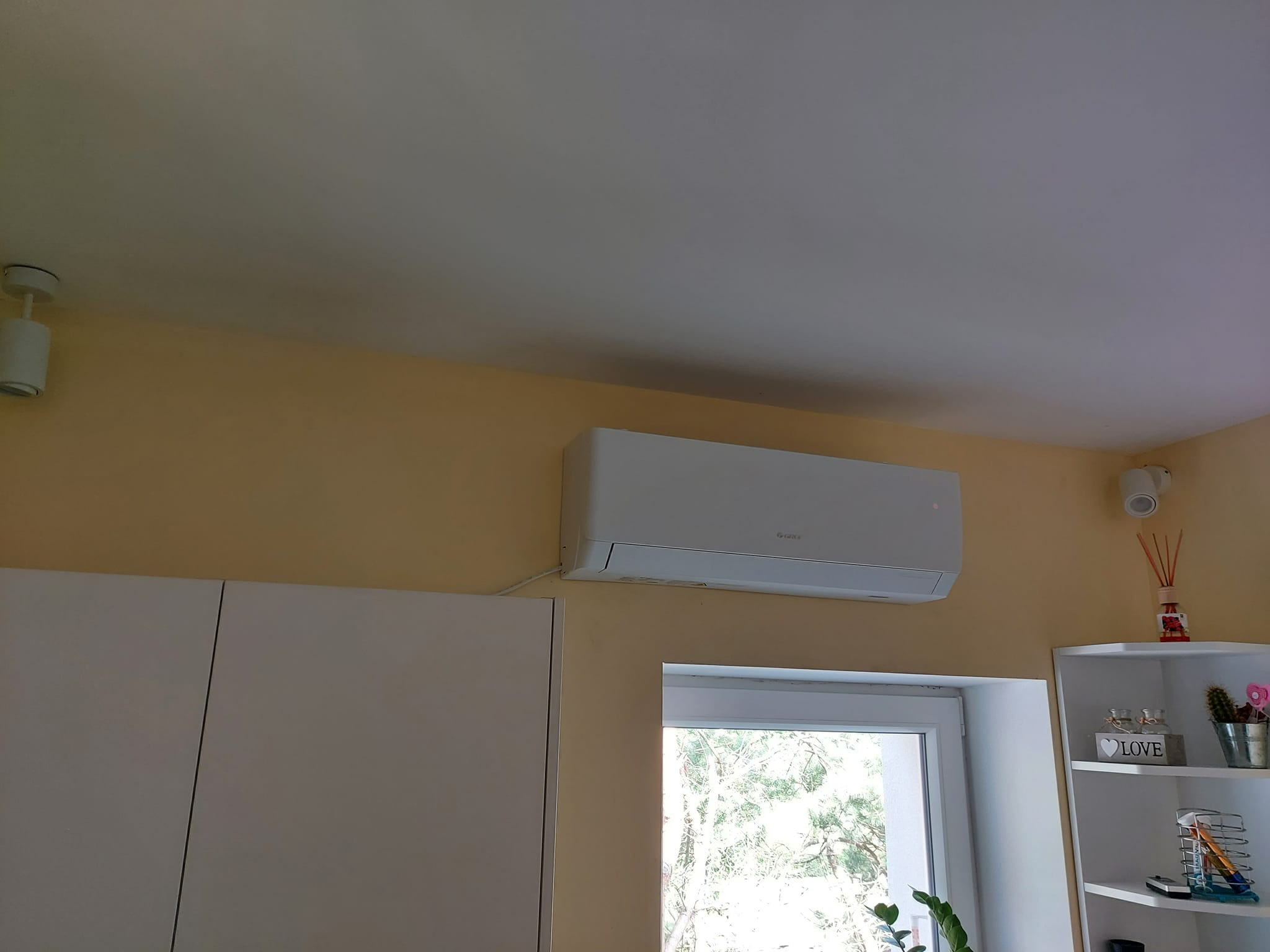 Biały klimatyzator marki Gree zamontowany na żółtej ścianie nad oknem w pokoju. Obok widoczny fragment białej szafy i rogu półki z dekoracjami.