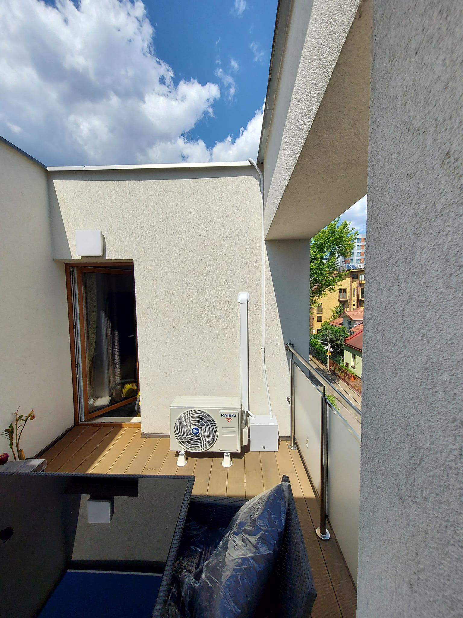 Jednostka zewnętrzna klimatyzatora marki Kaisai zamontowana na balkonie, widoczna instalacja elektryczna i rura odprowadzająca skropliny, w tle błękitne niebo z chmurami.
