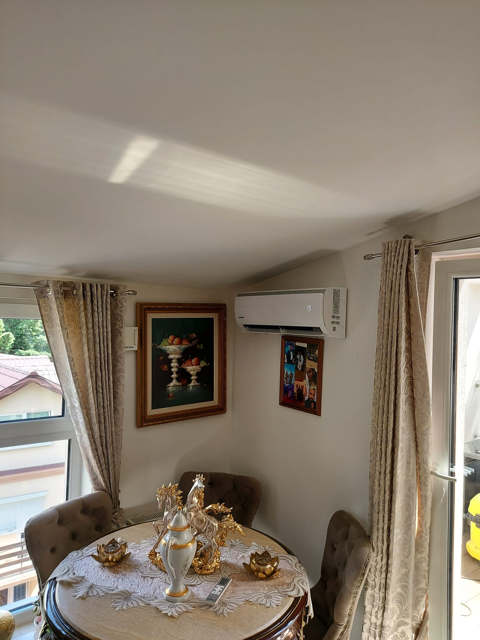 Elegancka jadalnia z zamontowaną klimatyzacją ścienną, widoczny stół z dekoracyjnym serwisem, obrazy na ścianie i zasłony przy oknie.