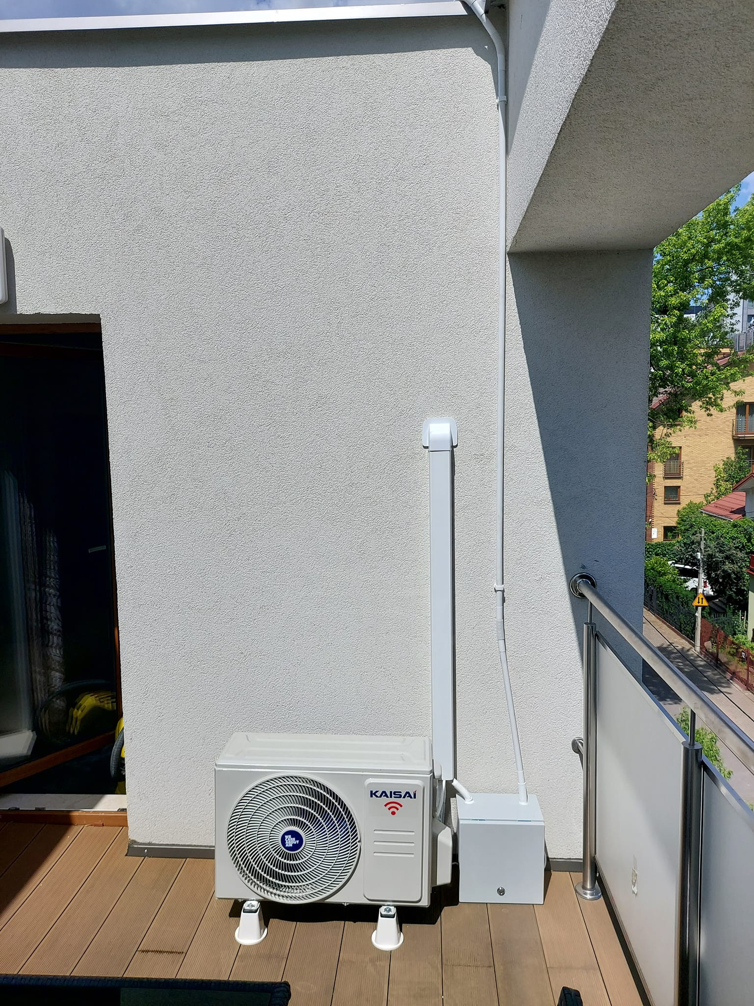 Biała jednostka zewnętrzna klimatyzacji marki Kaisai, zamontowana na balkonie z drewnianą podłogą, z widocznymi białymi rurami instalacyjnymi przymocowanymi do ściany budynku.