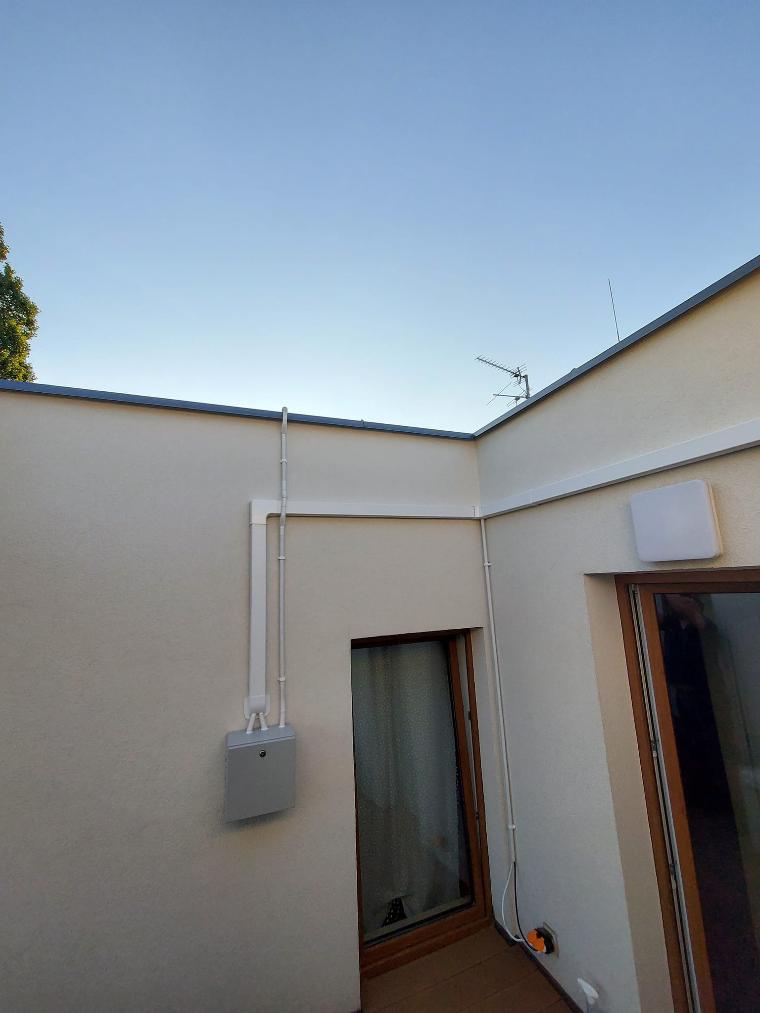 Zewnętrzna instalacja rekuperacji z szarą skrzynką przyłączeniową na elewacji budynku, widoczne białe rury prowadzone wzdłuż ściany i pod dachem, antena telewizyjna na dachu, jasne niebo w tle.