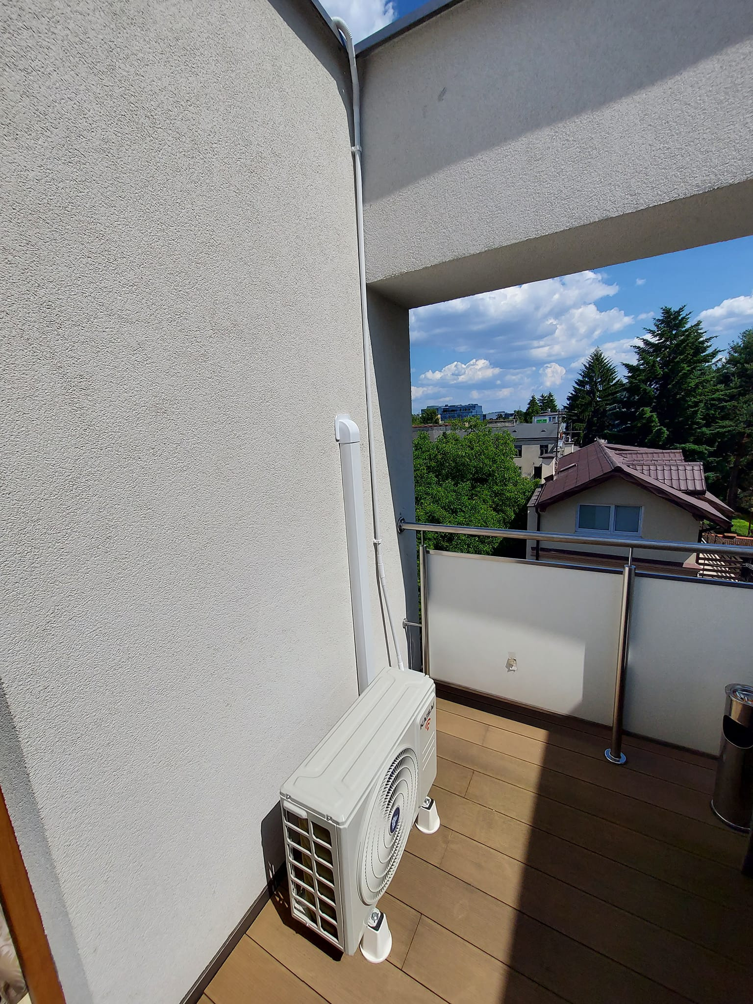 Zewnętrzna jednostka klimatyzacji marki Kaisai zamontowana na drewnianym balkonie, widok na okoliczną zabudowę i błękitne niebo.