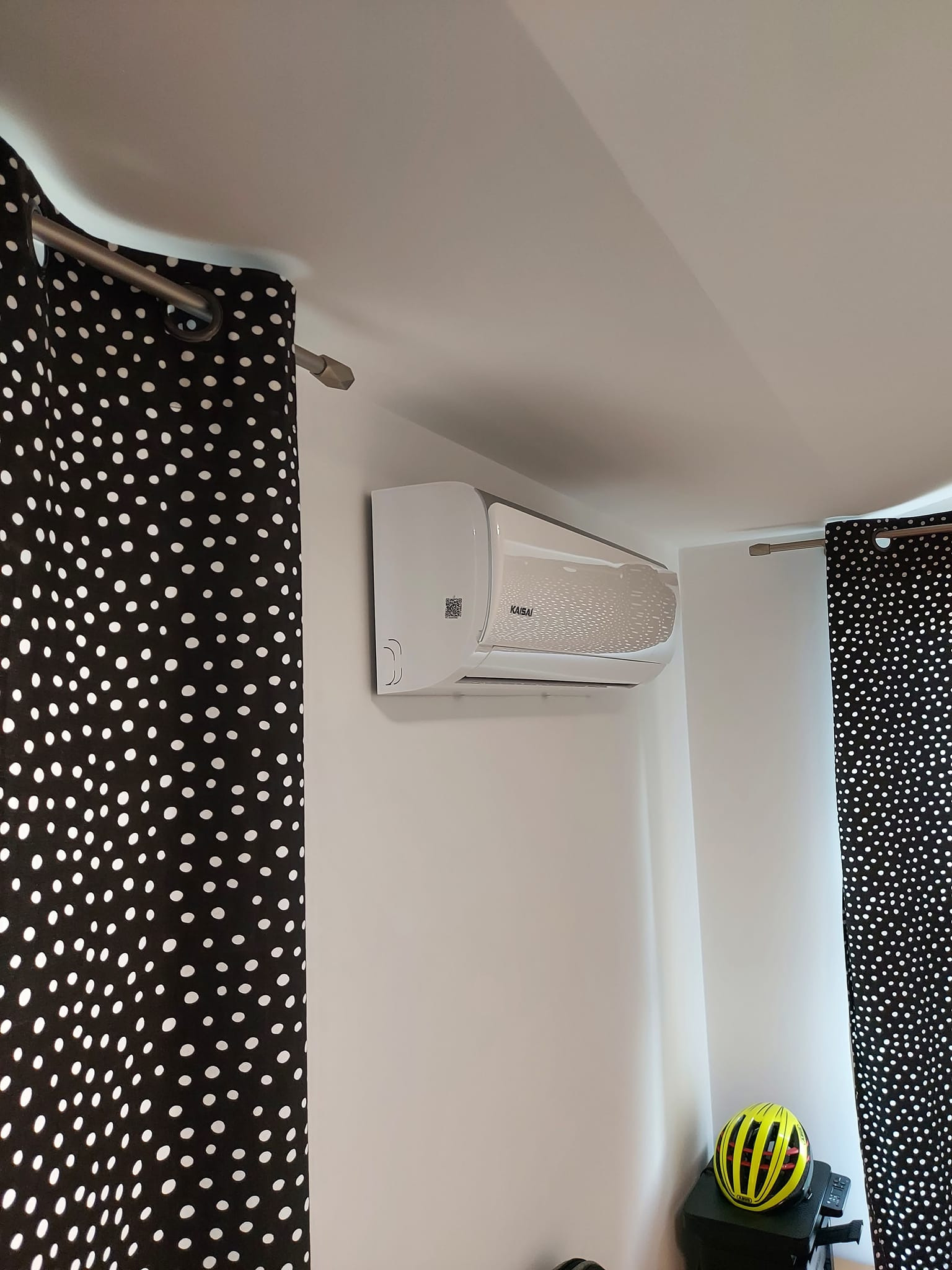 Biała jednostka klimatyzacji Kaisai zamontowana na ścianie w pokoju, widoczna z perspektywy lekko od dołu, z zasłonami w czarno-białe kropki po bokach i żółtym kaskiem rowerowym na szafce w tle.