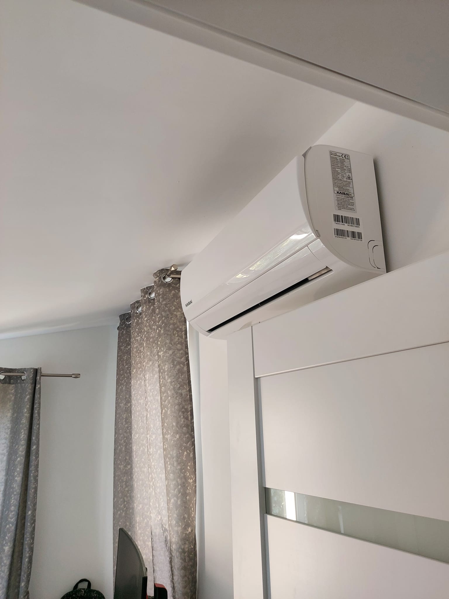 Biała jednostka klimatyzacji Kaisai zamontowana na ścianie nad szafą z przesuwnymi drzwiami i szarymi zasłonami w tle.