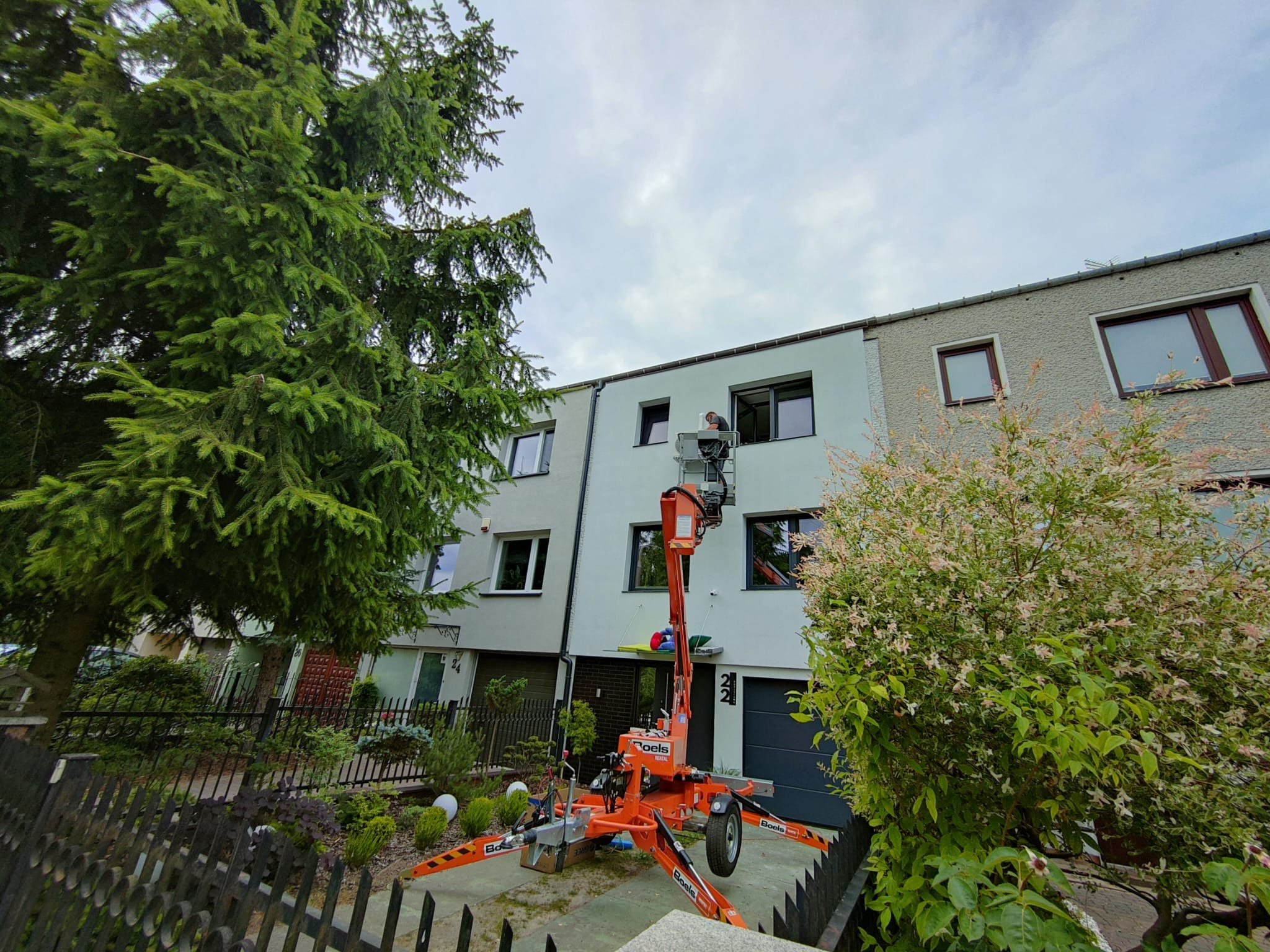 Pracownik na podnośniku koszowym przed budynkiem mieszkalnym, prawdopodobnie montujący lub serwisujący urządzenie na elewacji.