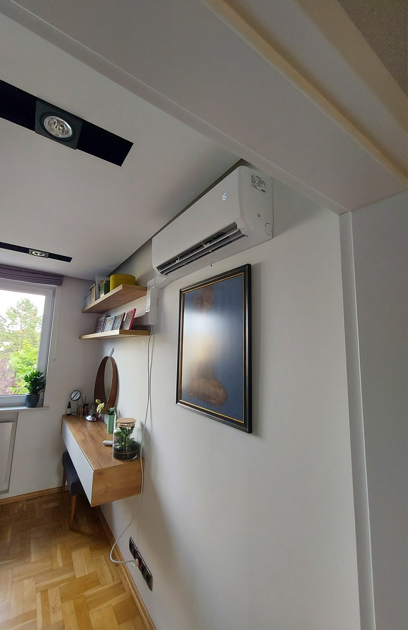 Biała jednostka wewnętrzna klimatyzatora zamontowana na ścianie w jasnym pomieszczeniu, obok wisi obraz w ciemnej ramie, widoczna toaletka z lustrem i półki na ścianie.