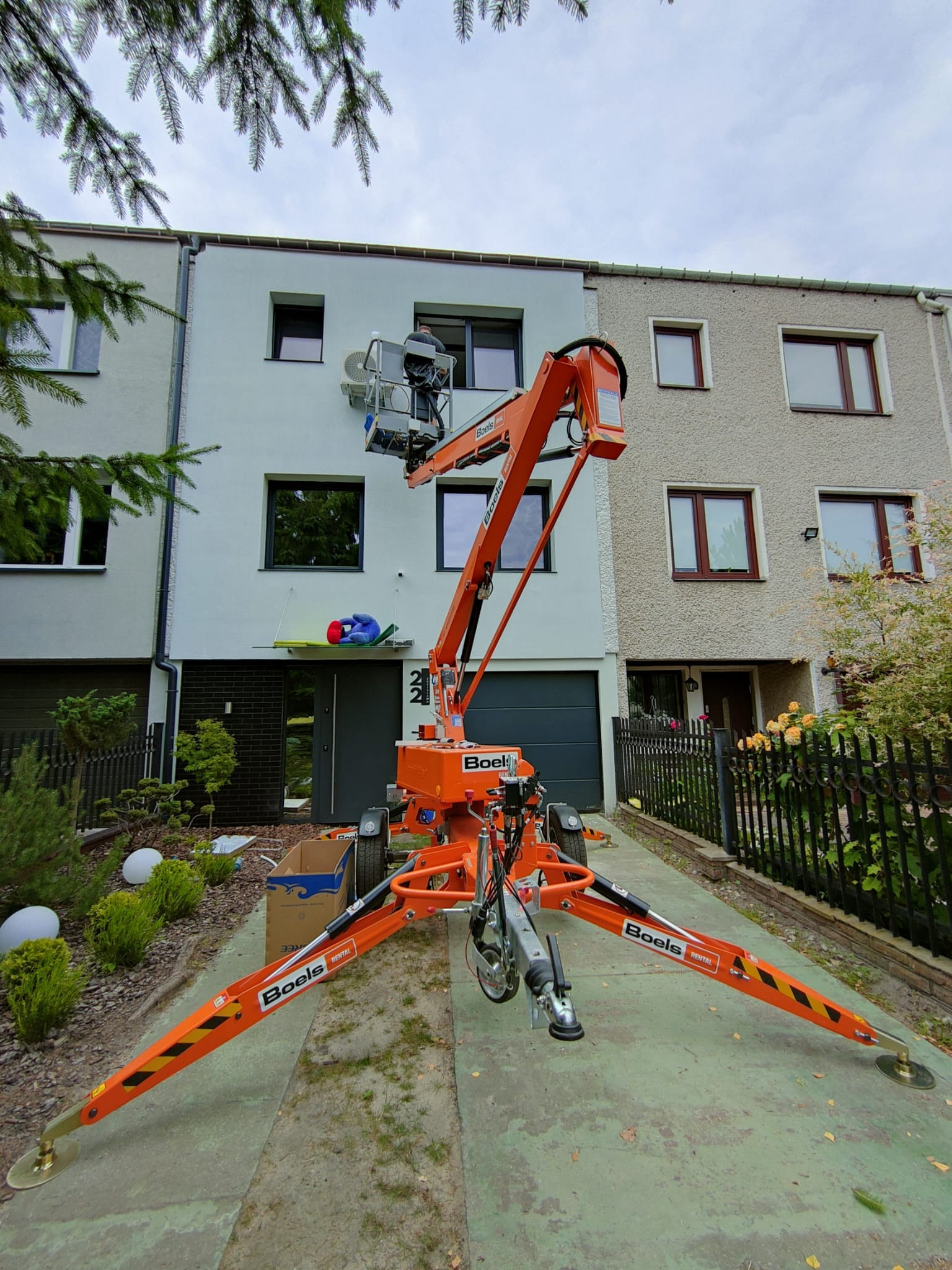 Montaż zewnętrznej jednostki klimatyzacji na elewacji domu z użyciem podnośnika koszowego firmy Boels, widok z dołu na pracownika w koszu na tle okna.