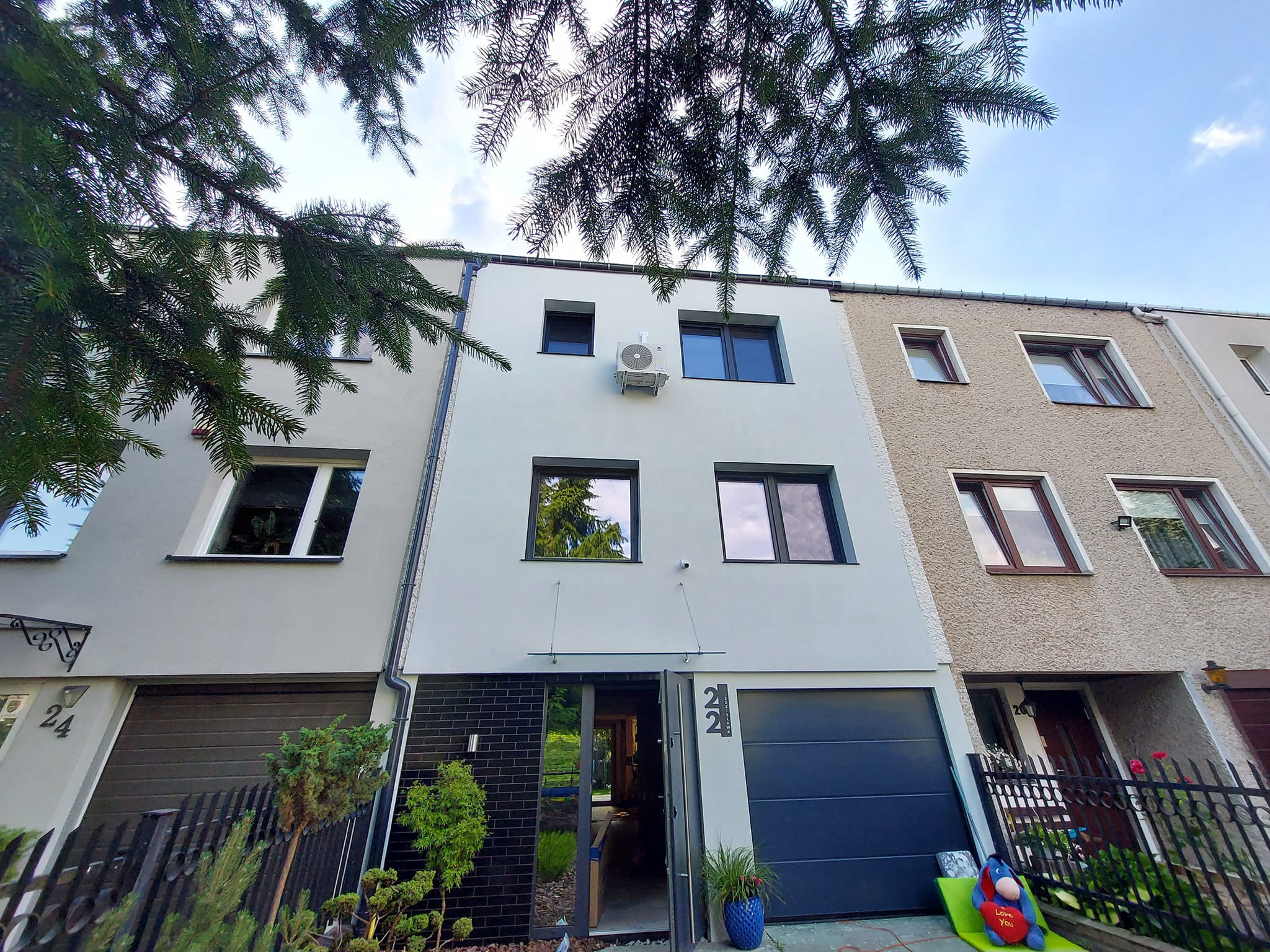 Elewacja nowoczesnego szeregowca z zamontowaną klimatyzacją na wysokości pierwszego piętra, widok z dołu przez gałęzie drzewa.