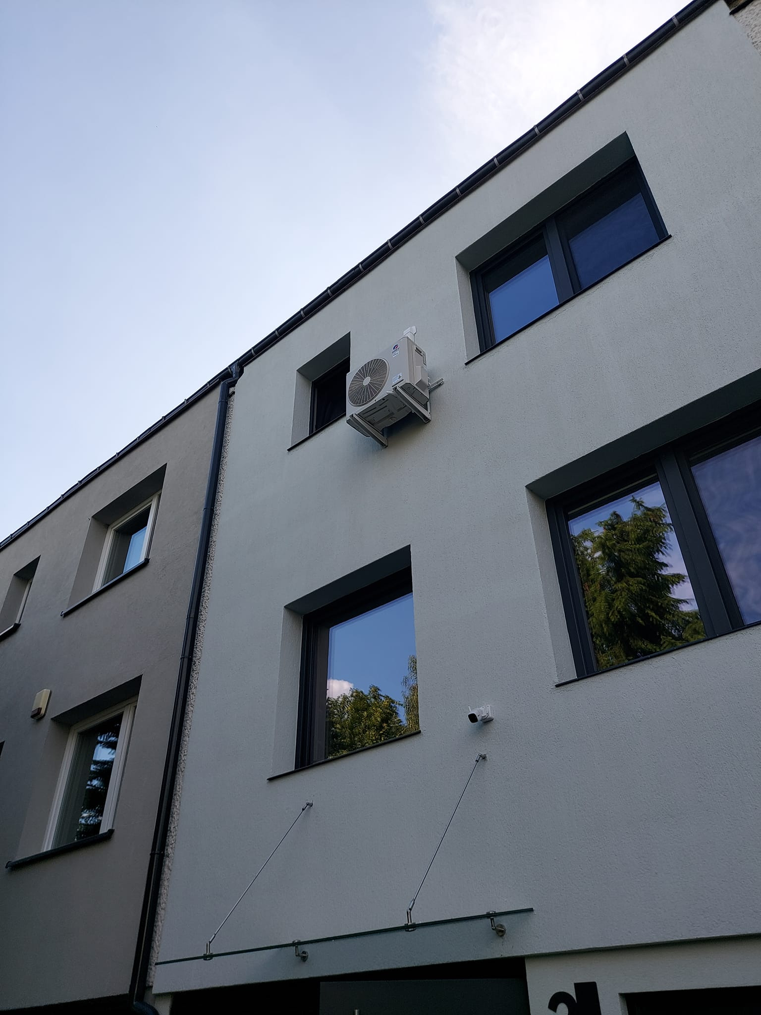 Zewnętrzna jednostka klimatyzacji zamontowana na elewacji dwukondygnacyjnego budynku z ciemnymi oknami, widok z dołu pod kątem.