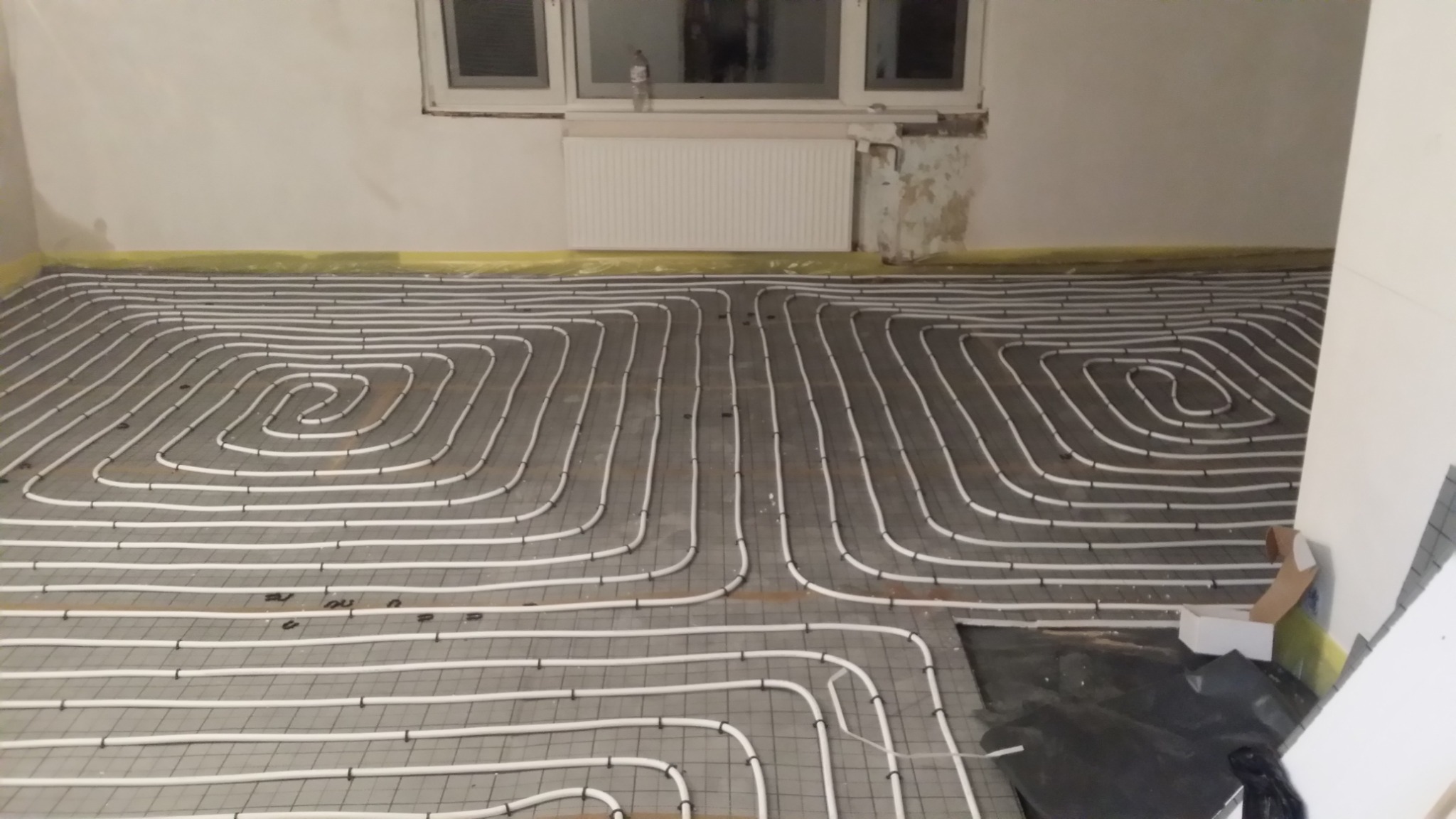 Układ rur ogrzewania podłogowego na siatce montażowej, widoczne białe rury w kształcie meandrów, żółta taśma przy ścianie, fragment grzejnika pod oknem, przygotowanie do wylewki.