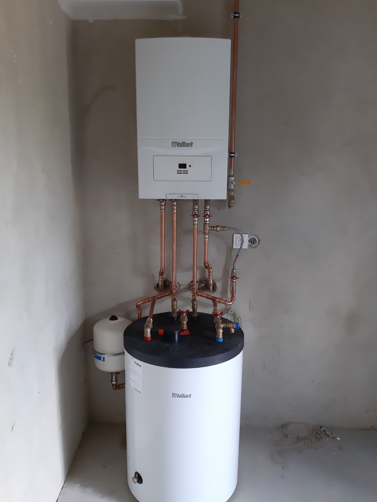 Biały piec grzewczy Vaillant zamontowany na ścianie z widocznymi miedzianymi rurami i białym zasobnikiem wody z czarnym blatem, w surowym pomieszczeniu.