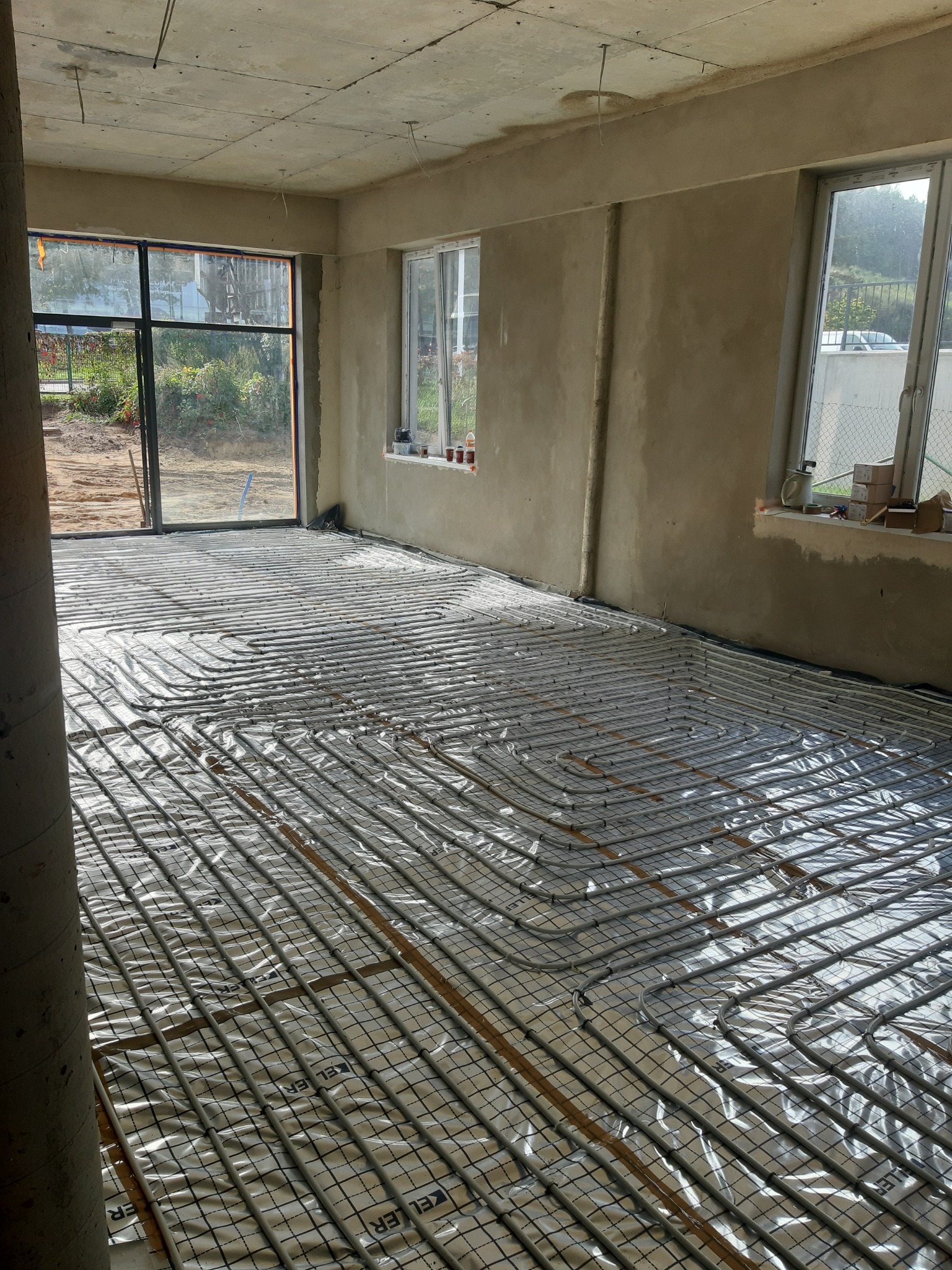 Instalacja ogrzewania podłogowego w surowym wnętrzu, rury grzewcze rozłożone na folii z nadrukiem, widok na okno i niezagospodarowany teren na zewnątrz.