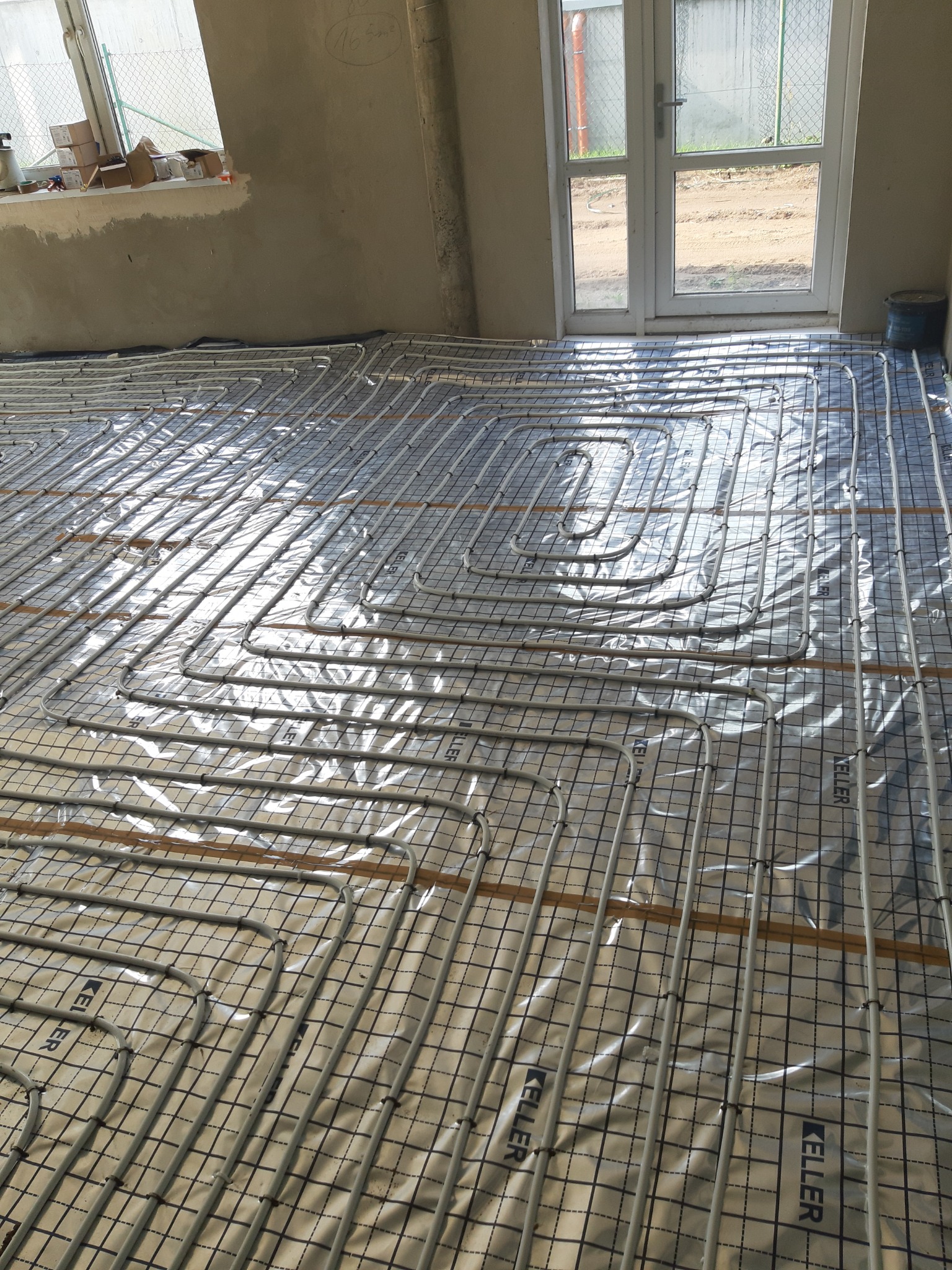 Szare rury ogrzewania podłogowego ułożone na folii z nadrukiem kratki i logo Keller, widoczne w surowym wnętrzu z oknem i drzwiami balkonowymi.