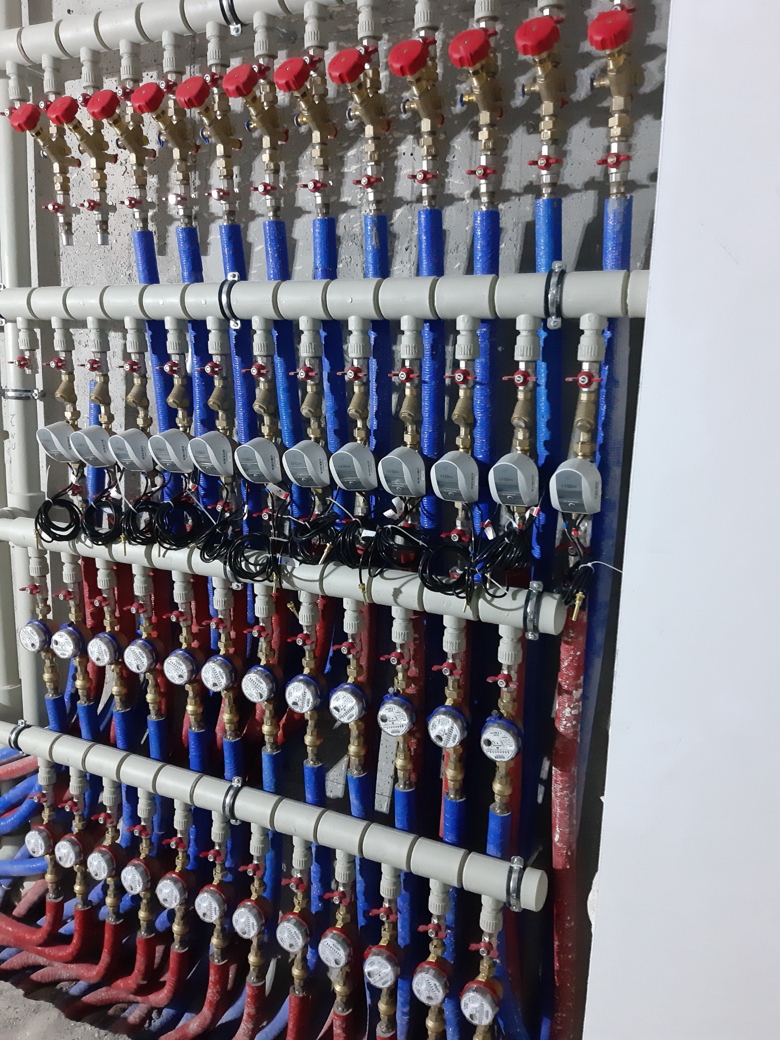 Rozdzielacz ogrzewania podłogowego z licznymi zaworami, rurami w izolacji niebieskiej i czerwonej oraz białymi licznikami ciepła, zamontowany przy szarej ścianie.
