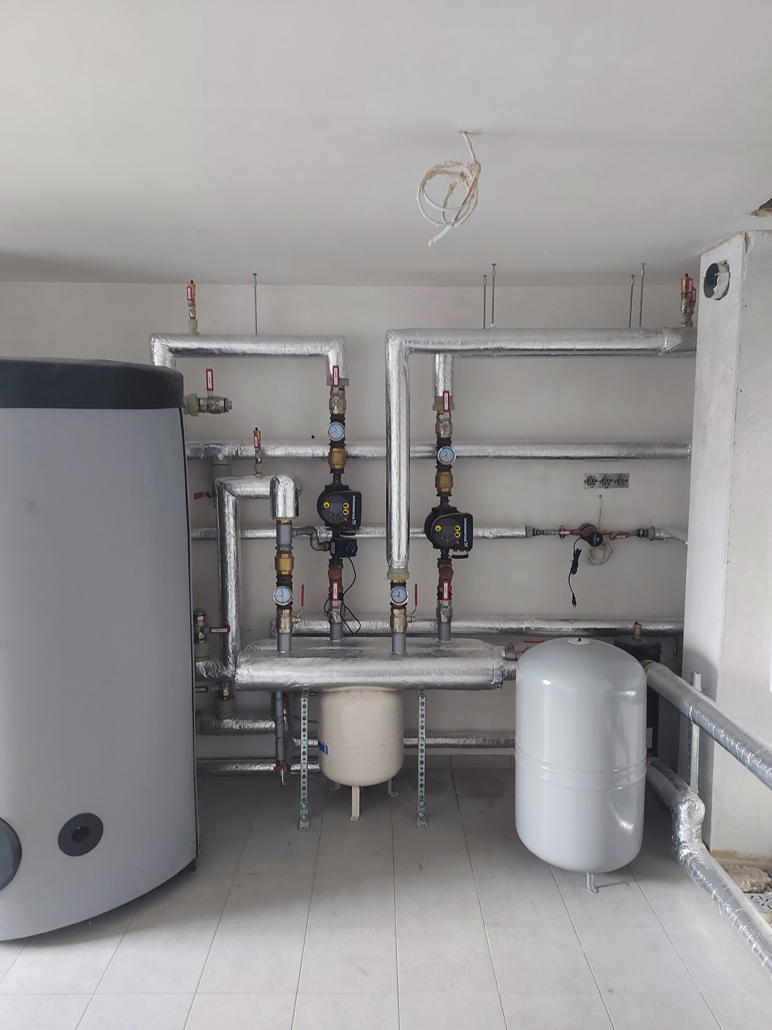 Kotłownia z szarym zbiornikiem buforowym, rurami w srebrnej izolacji, pompami obiegowymi i białymi zbiornikami wyrównawczymi, widok na instalację po montażu.