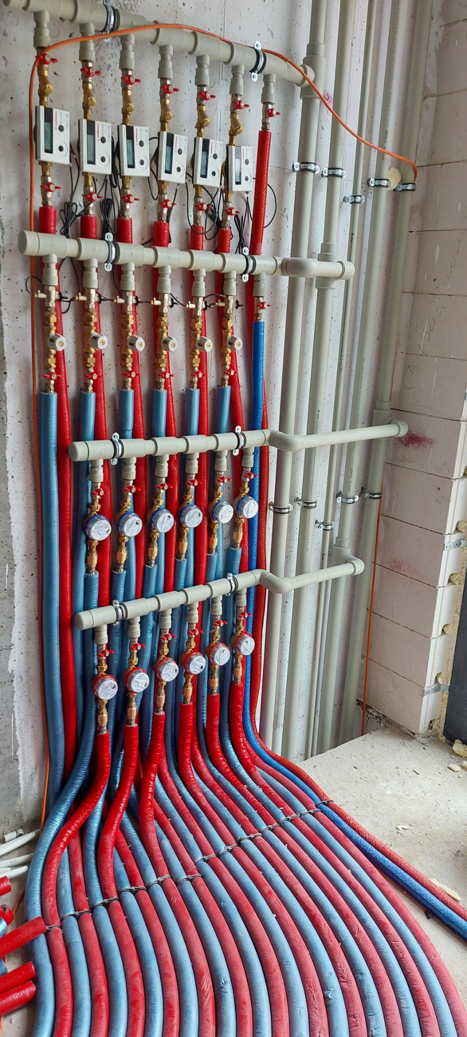 Rozdzielacz ogrzewania podłogowego z licznikami ciepła i rurami w izolacji czerwonej i niebieskiej, zamontowany na ścianie z bloczków betonu komórkowego.