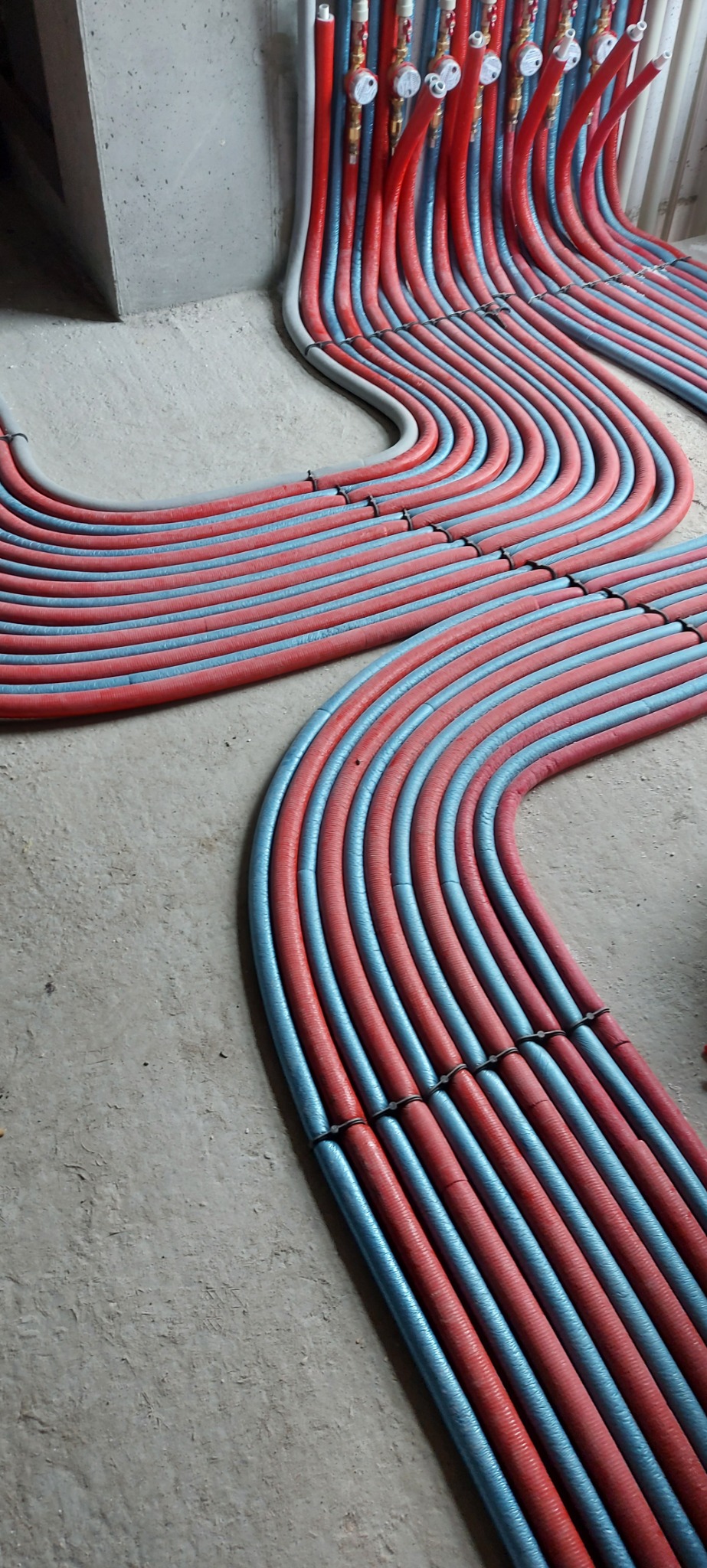 Rozłożone na podłodze rury instalacji grzewczej w kolorach czerwonym i niebieskim, ułożone równolegle i przymocowane czarnymi zaciskami, z widocznym fragmentem betonowej ściany w tle.