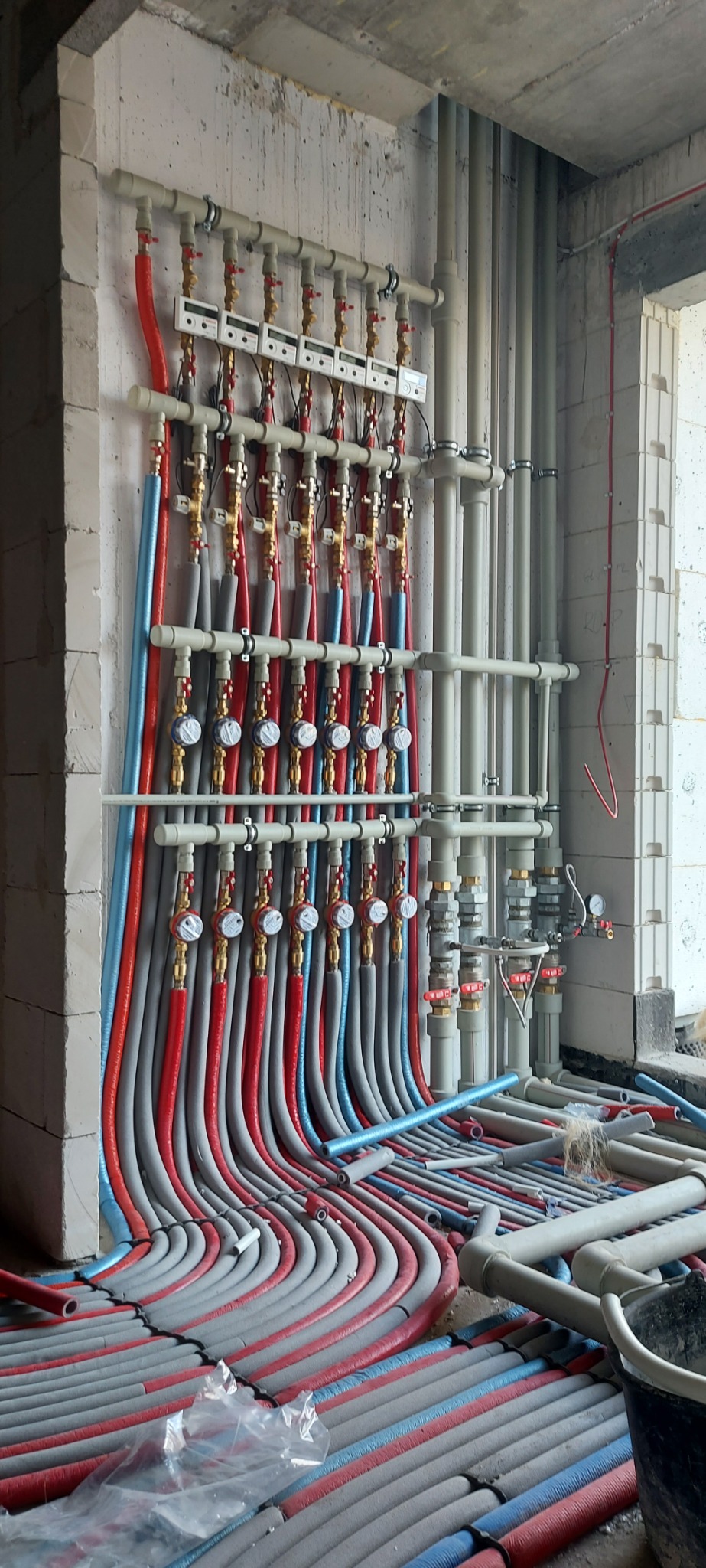 Rozdzielacz ogrzewania podłogowego z rurami w izolacji czerwono-szarej i niebiesko-szarej, zamontowany na ścianie z bloków betonowych w surowym stanie.