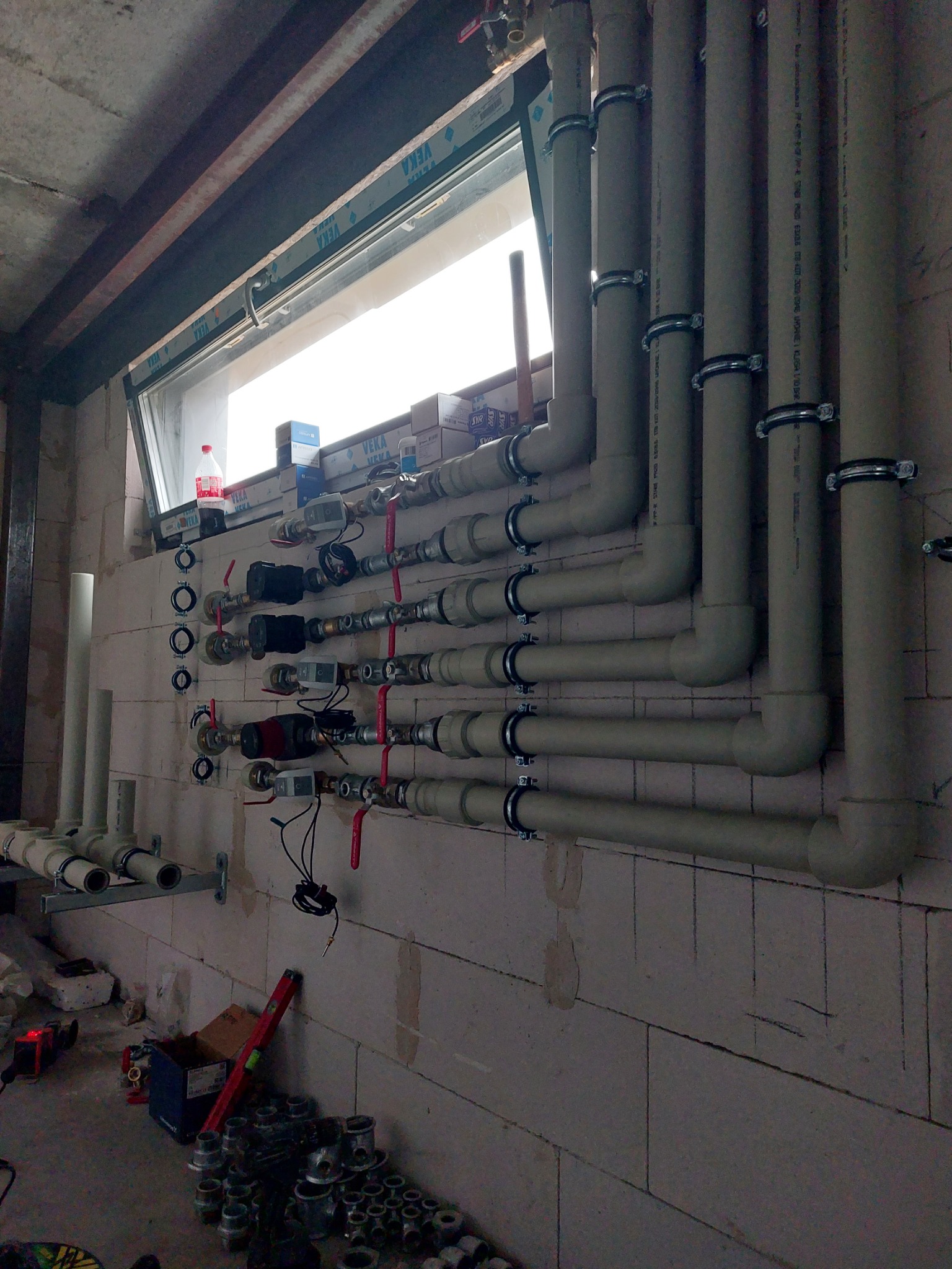 Instalacja rur grzewczych i zaworów przy oknie w surowym stanie, widoczne liczne rury pionowe i poziome, zawory z czerwonymi uchwytami, przewody elektryczne, oraz narzędzia i materiały budowlane...