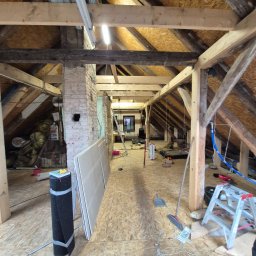 Klimas Remonty - Adaptacja poddasza: widok na odsłoniętą konstrukcję dachu z drewnianymi belkami, przygotowane płyty gipsowe i narzędzia. W tle widoczny komin z cegły.