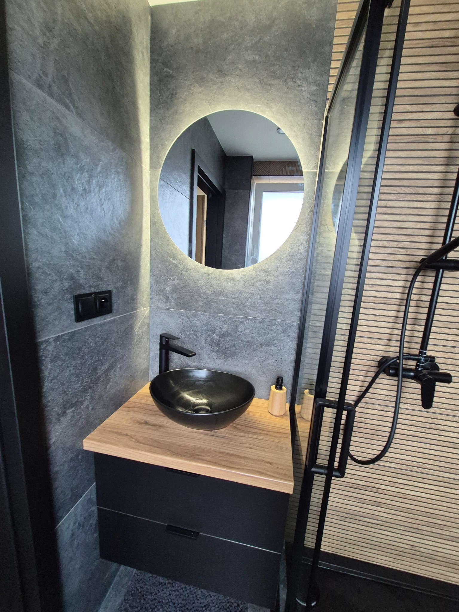 Nowoczesna łazienka: szare płytki imitujące kamień, okrągłe lustro z podświetleniem, czarna umywalka nablatowa i kabina prysznicowa z czarną ramą.