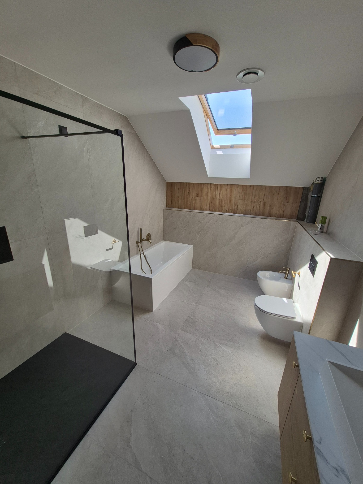 Stylowa łazienka na poddaszu z kabiną typu walk-in, wanną, toaletą, bidetem i umywalką. Jasne płytki imitujące kamień, złota armatura i drewniane akcenty.