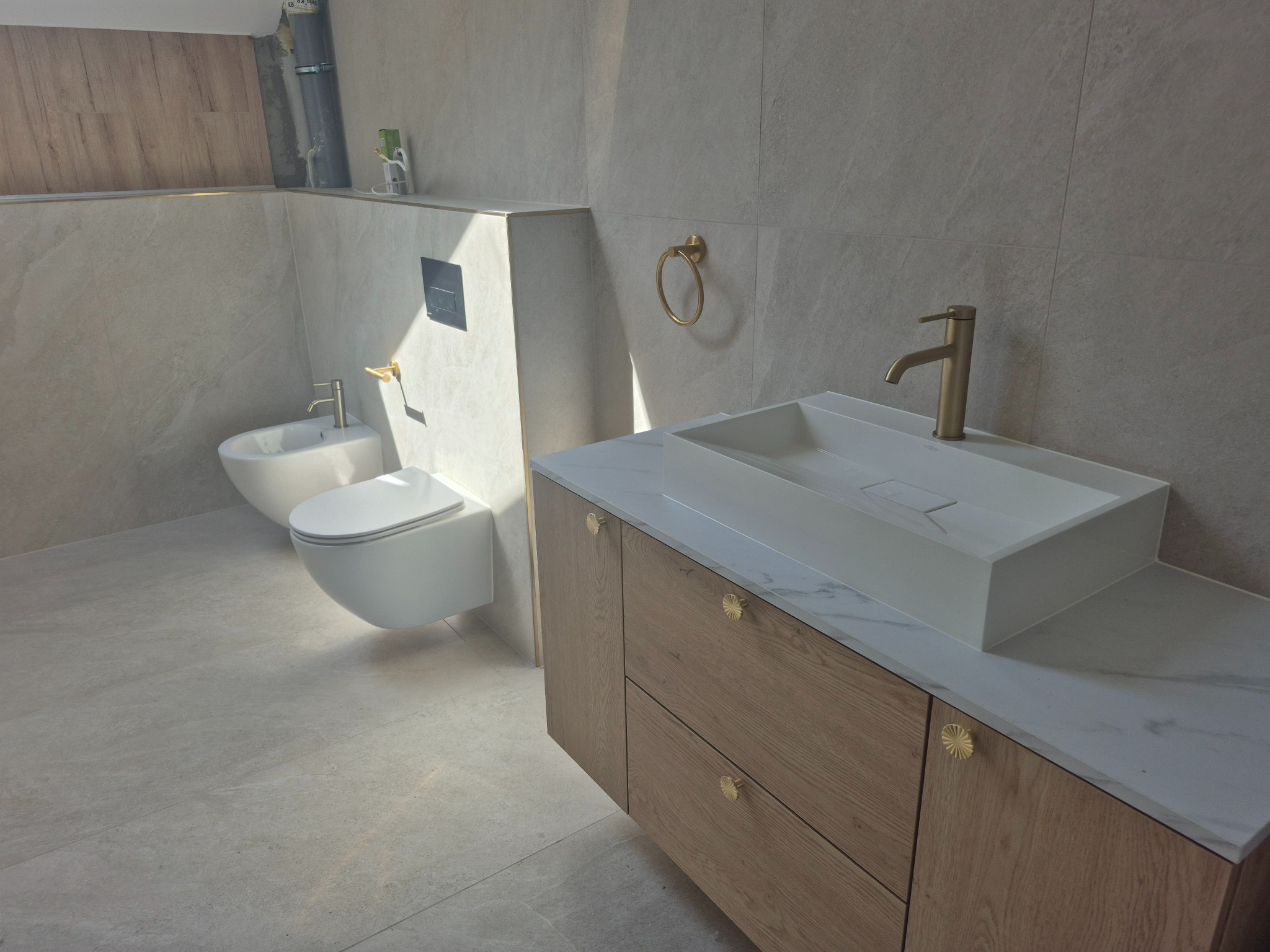 Nowoczesna łazienka z beżowymi płytkami, białą ceramiką i drewnianą szafką z umywalką. Bateria i akcesoria w kolorze złotym, dodające elegancji.