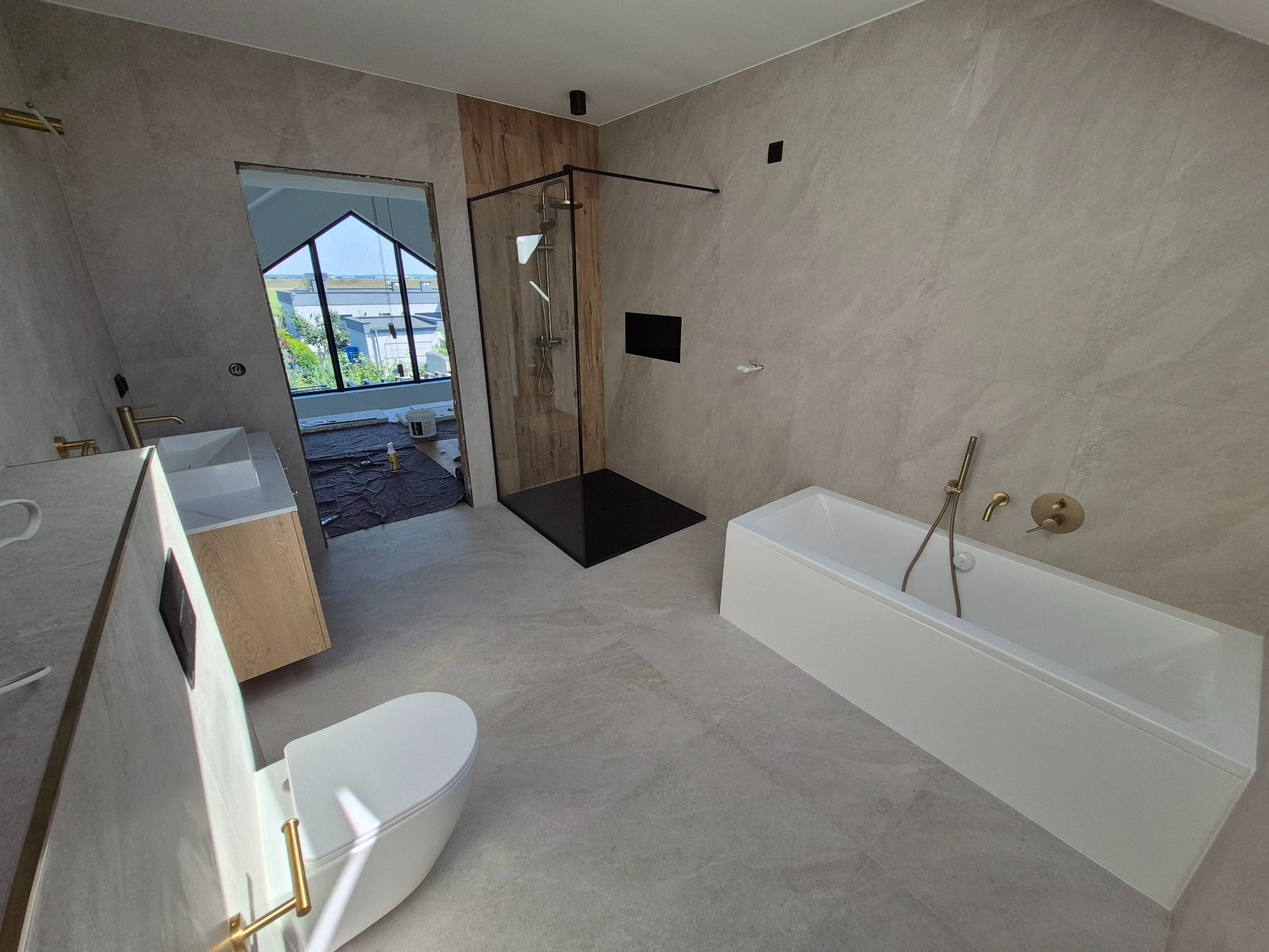 Nowoczesna łazienka w odcieniach szarości z elementami drewna, kabiną prysznicową typu walk-in i wolnostojącą wanną. Widoczne złote detale armatury.