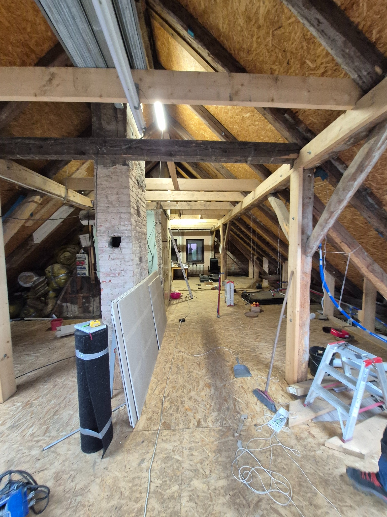 Adaptacja poddasza: widok na odsłoniętą konstrukcję dachu z drewnianymi belkami, przygotowane płyty gipsowe i narzędzia. W tle widoczny komin z cegły.