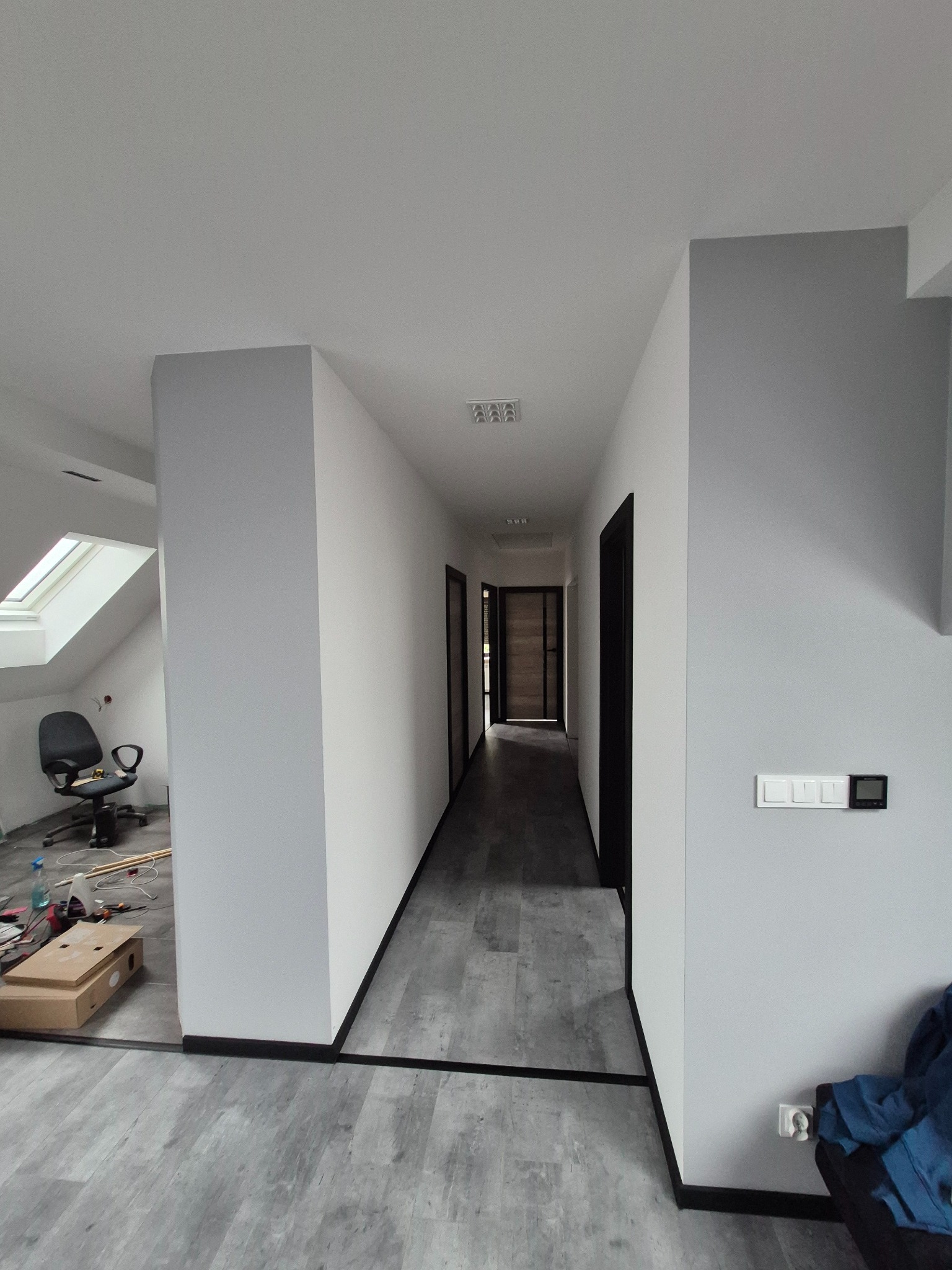 Korytarz w szarościach po remoncie: szare panele, białe ściany z czarnymi listwami przypodłogowymi i ościeżnicami. W tle pokój z oknem dachowym i krzesłem biurowym.