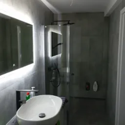 Nowoczesna łazienka z szarymi płytkami, kabiną prysznicową typu walk-in z czarną armaturą i umywalką nablatową z czarnym kranem, oświetlonym lustrem i widocznym fragmentem kabla wystającego z sufitu.