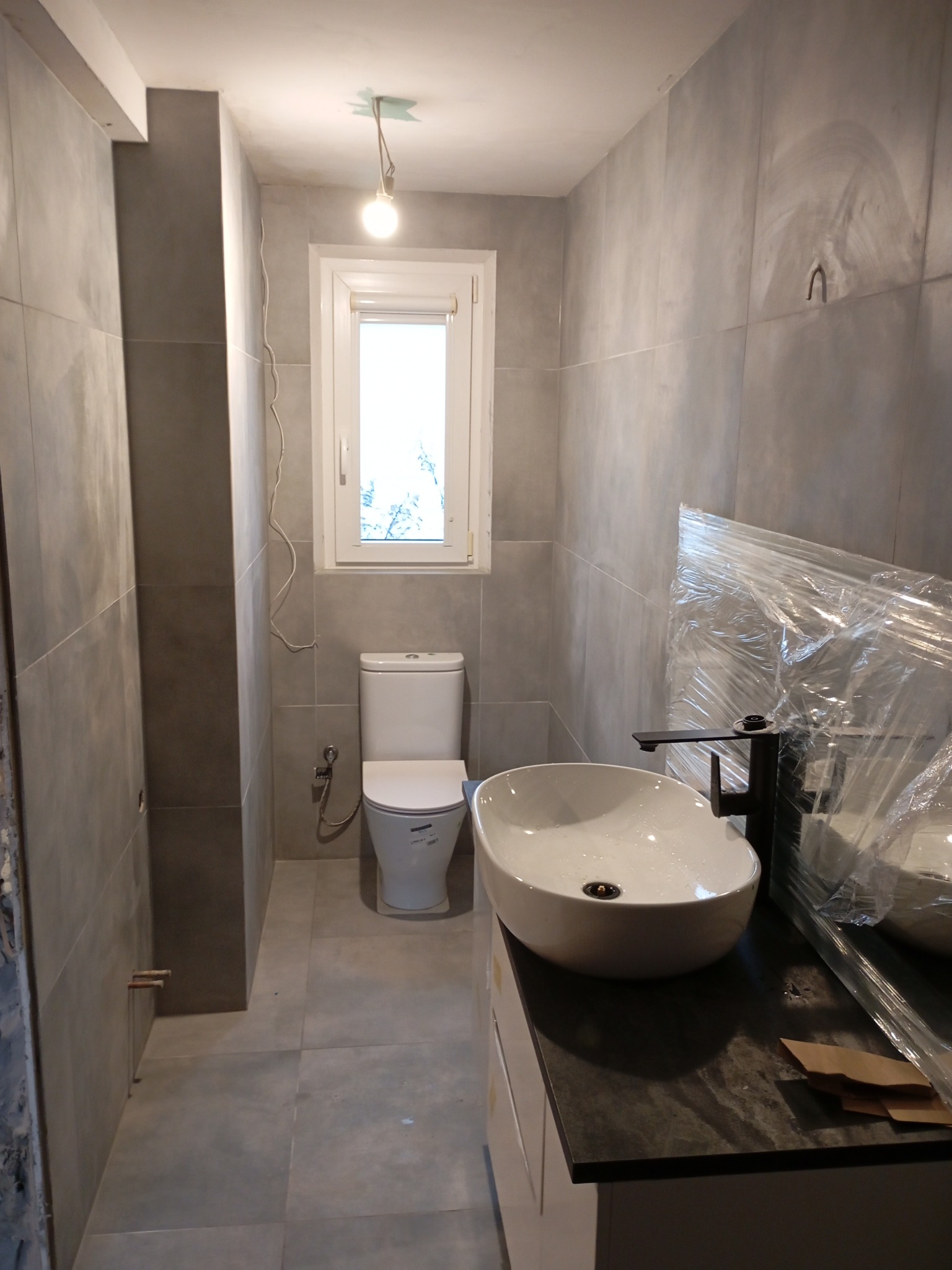 Nowoczesna łazienka w szarościach z białą ceramiką, umywalka nablatowa i toaleta kompaktowa, ściany wykończone płytkami imitującymi beton, lustro zabezpieczone folią ochronną.