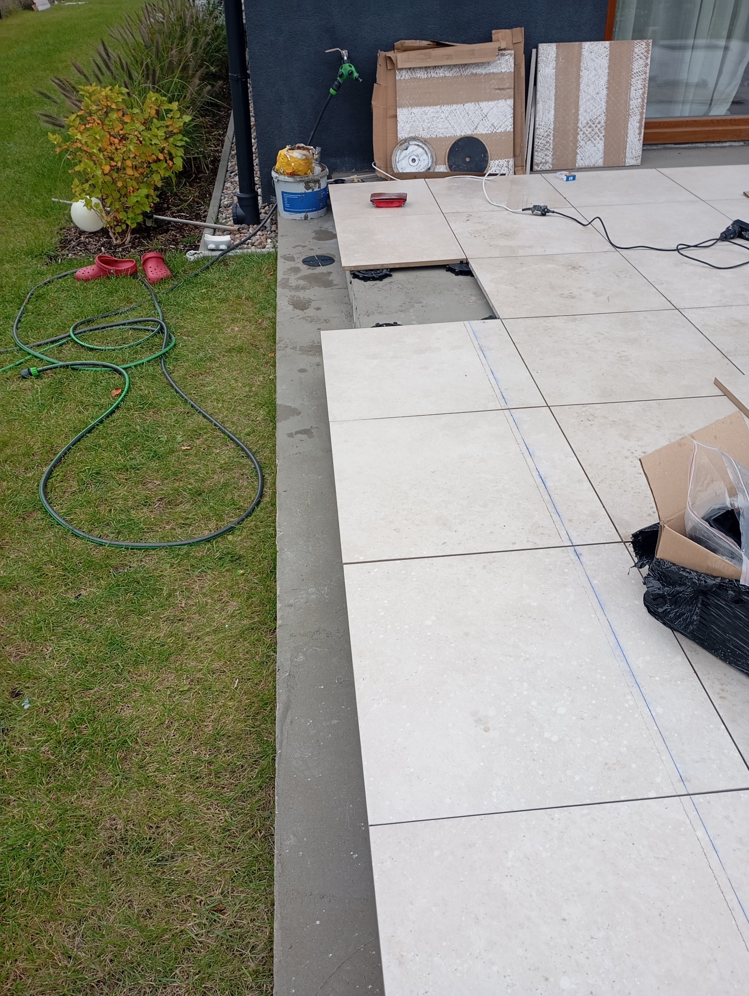 Układanie jasnych, kwadratowych płytek na tarasie, widoczne podkładki dystansowe, narzędzia glazurnicze i zaznaczone linie cięcia.