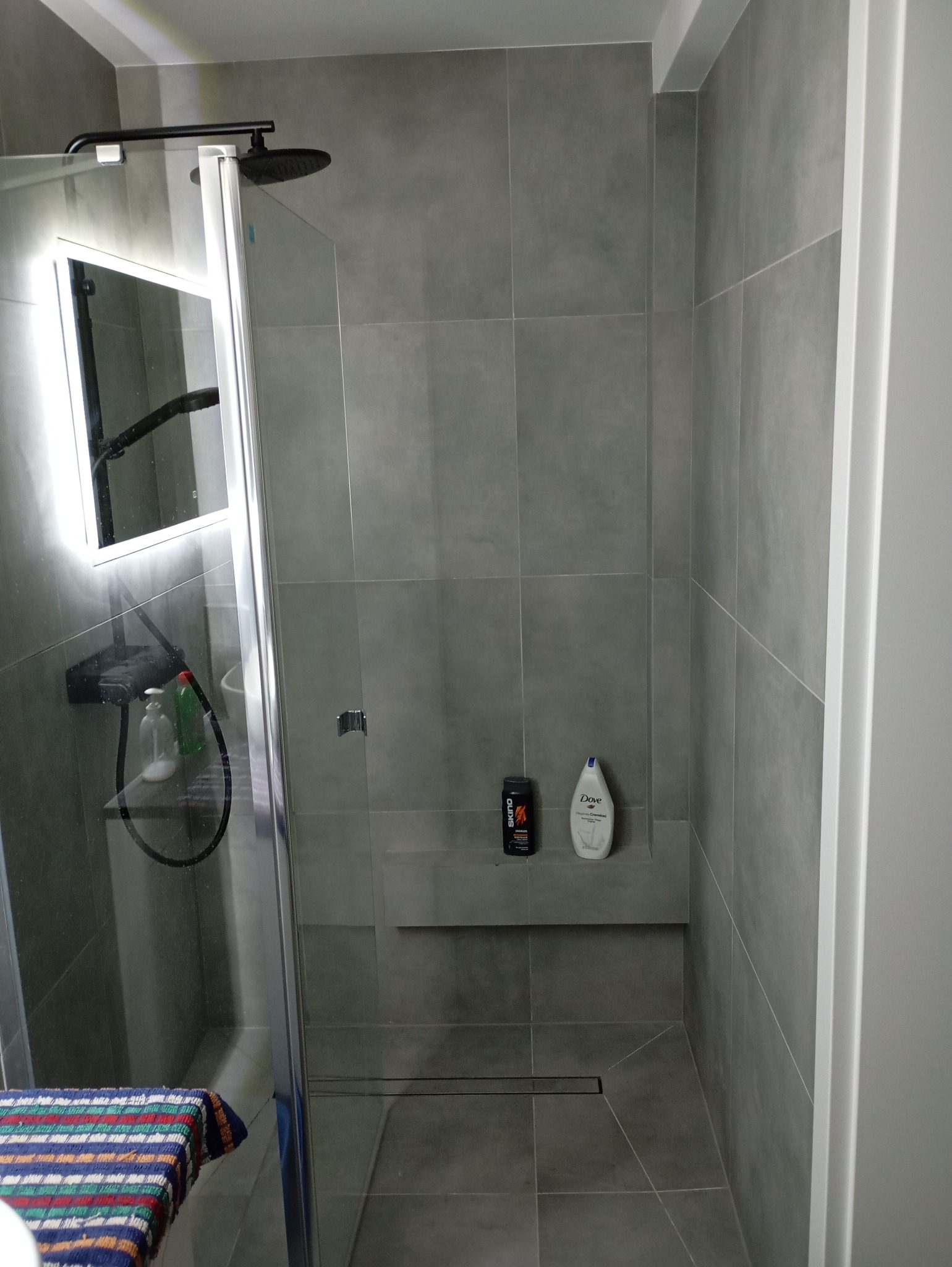 Nowoczesna kabina prysznicowa z deszczownicą i podświetlanym lustrem, szare płytki, półka z kosmetykami, odpływ liniowy.