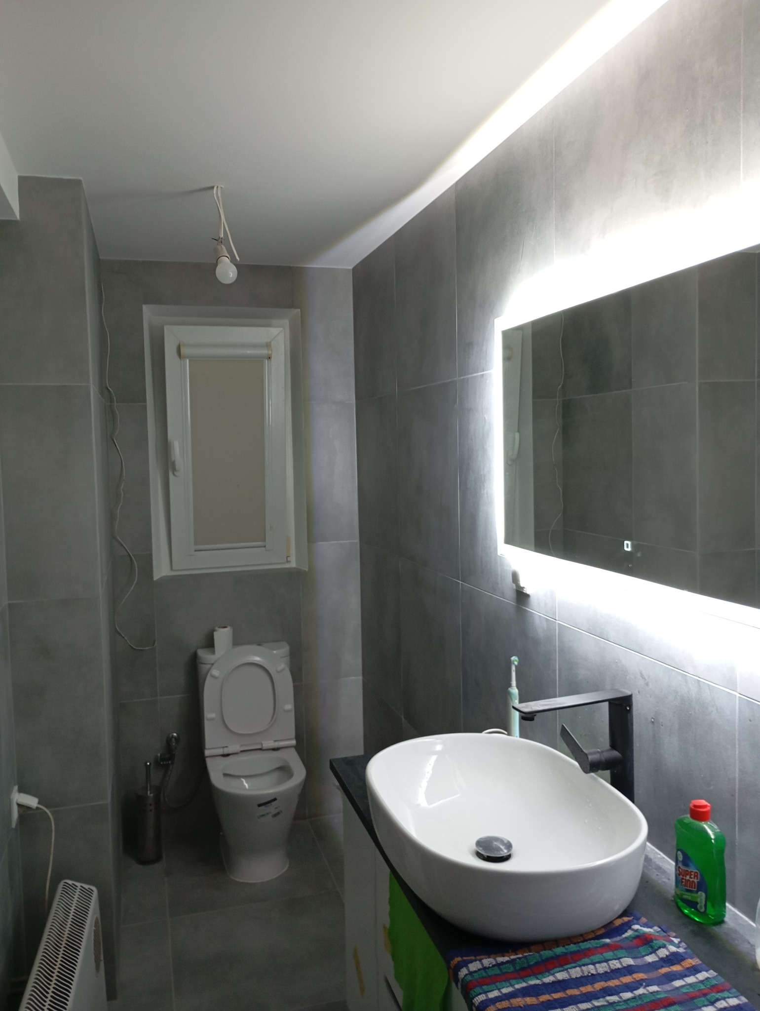 Szara łazienka po remoncie z podświetlanym lustrem, umywalką nablatową, toaletą i grzejnikiem.