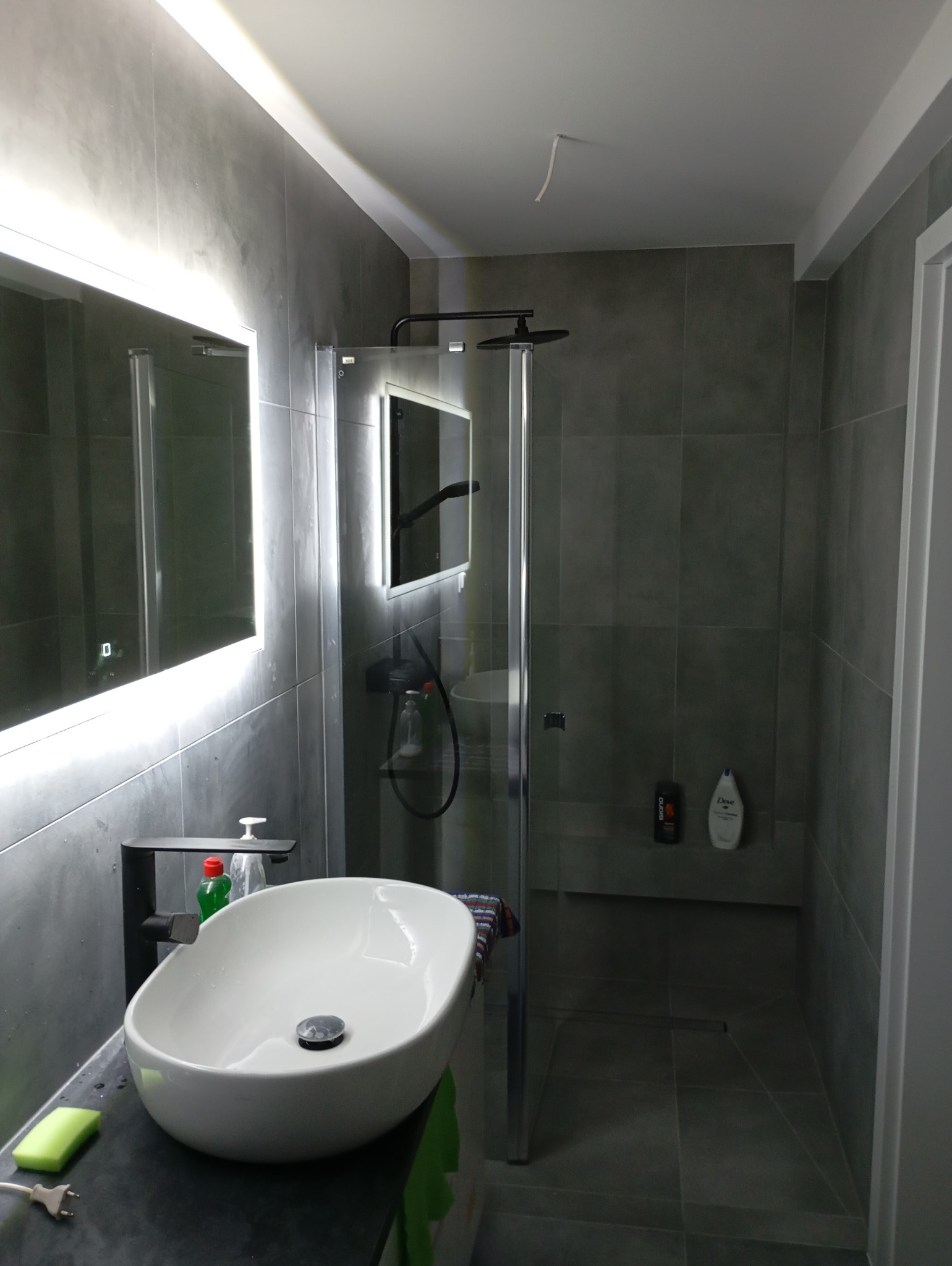 Nowoczesna łazienka z szarymi płytkami, kabiną prysznicową typu walk-in z czarną armaturą i umywalką nablatową z czarnym kranem, oświetlonym lustrem i widocznym fragmentem kabla wystającego z sufitu.