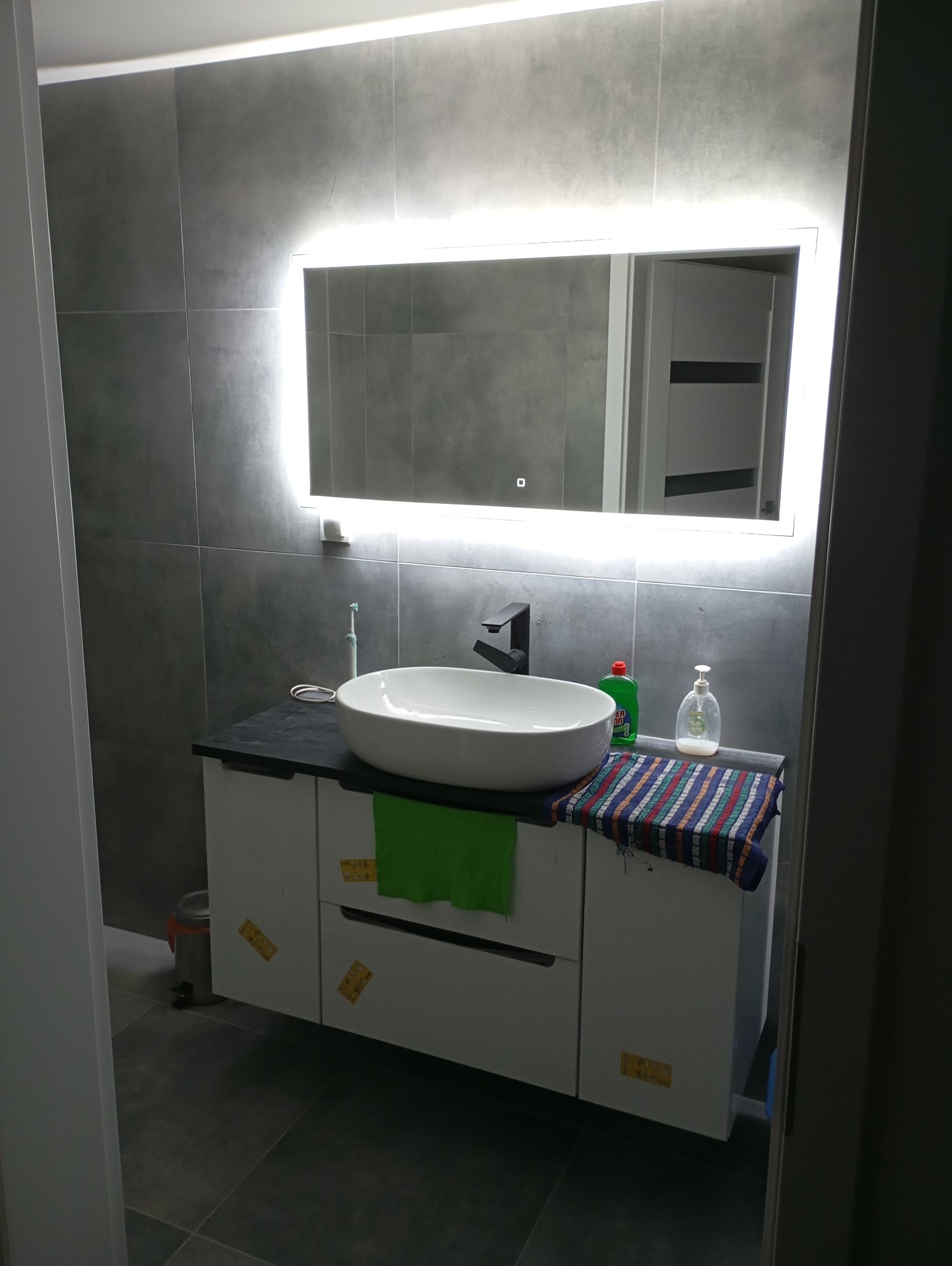 Nowoczesna łazienka z szarymi płytkami, podświetlanym lustrem, białą umywalką nablatową z czarną armaturą, zielonym ręcznikiem i kolorową szmatką na szafce pod umywalką.