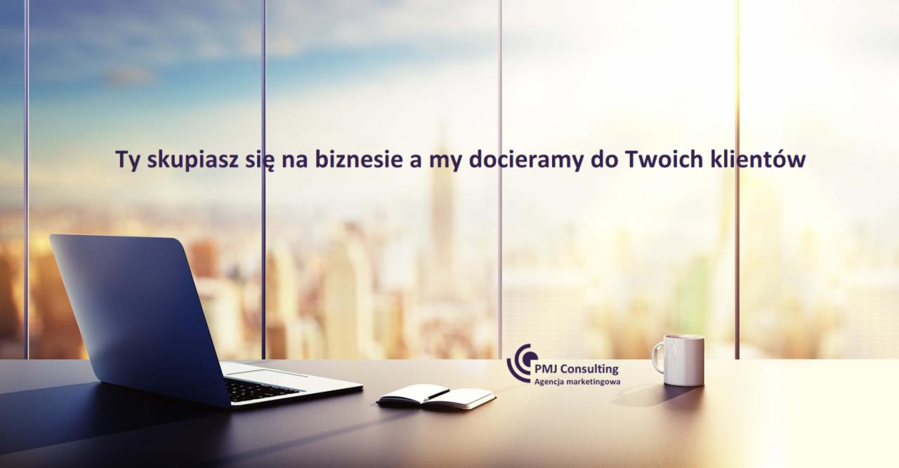 Laptop, notatnik i kubek na biurku z widokiem na miasto przez okno, nadrukowany slogan 'Ty skupiasz się na biznesie a my docieramy do Twoich klientów' oraz logo 'PMJ Consulting Agencja marketingowa'.