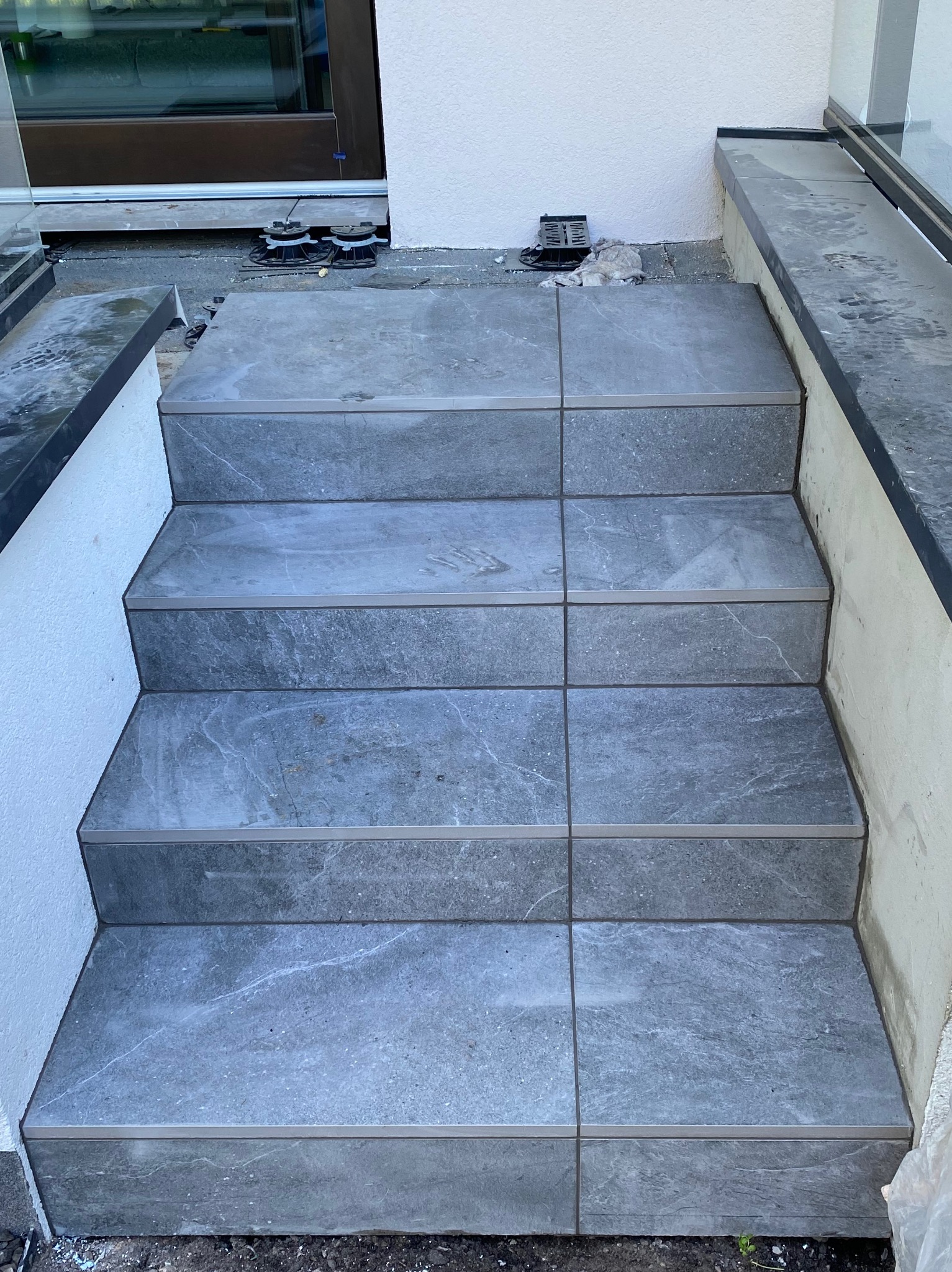 Schody zewnętrzne wyłożone szarymi, marmurkowymi płytkami, widoczne fugi i listwy wykończeniowe, w tle okno i fragment elewacji budynku.