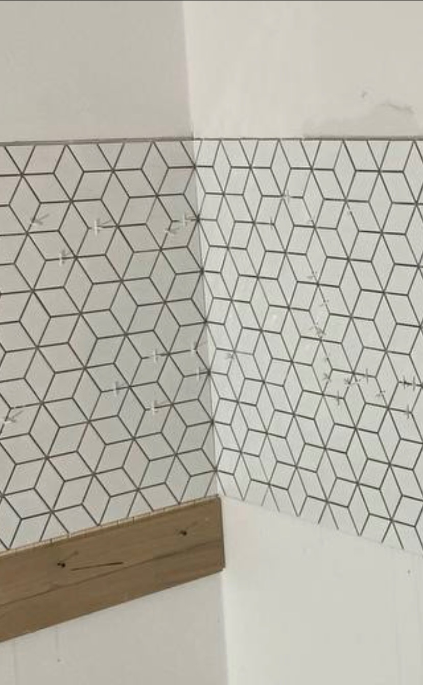 Układanie białych płytek z geometrycznym wzorem 3D w rogu pomieszczenia, z użyciem krzyżyków dystansowych i drewnianej listwy przypodłogowej.