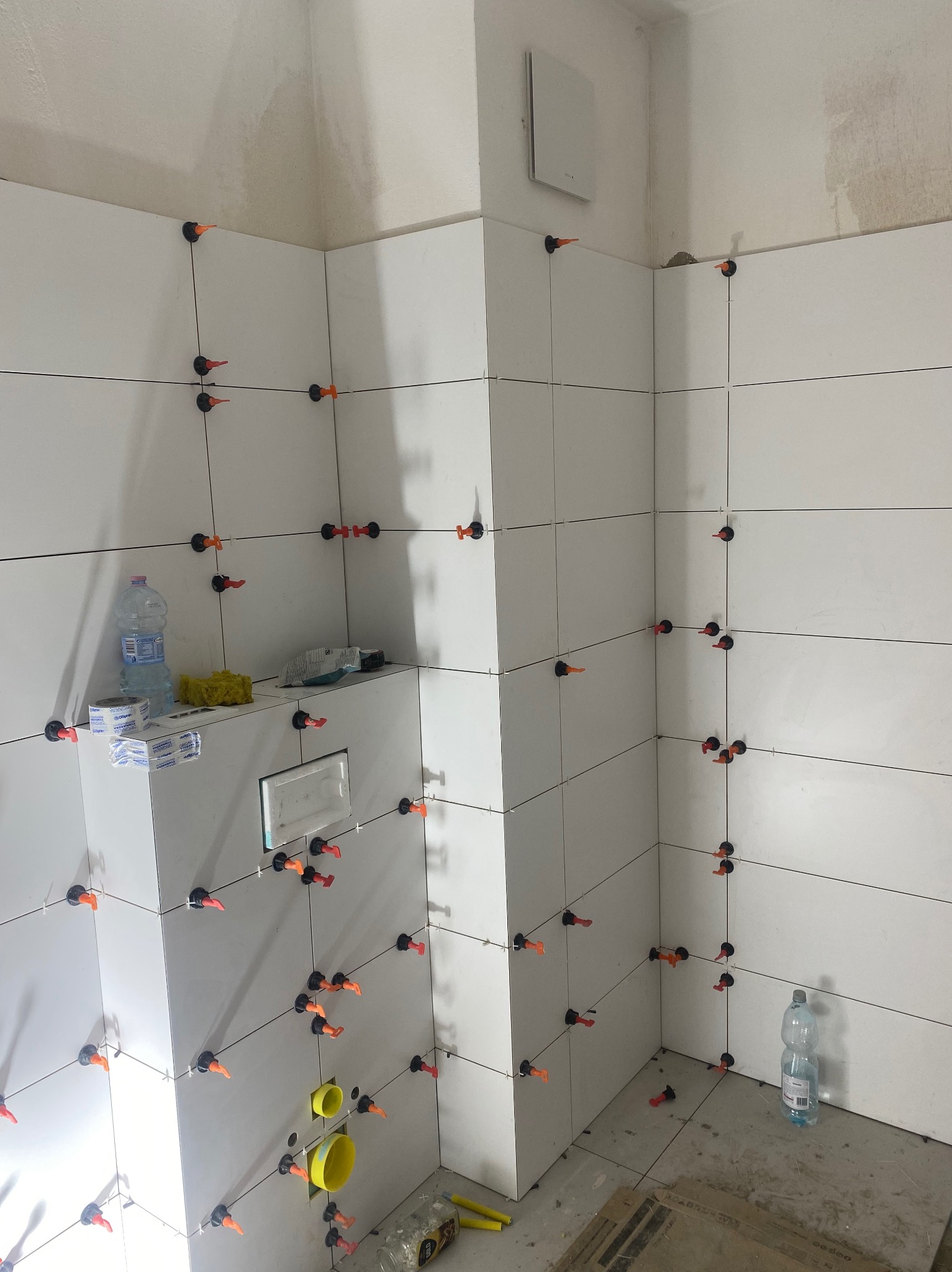 Świeżo położone, białe, prostokątne płytki w łazience, z widocznymi systemami poziomowania płytek (czarne podstawy i pomarańczowe kliny), żółte rury kanalizacyjne wystają ze ściany, butelka wody...