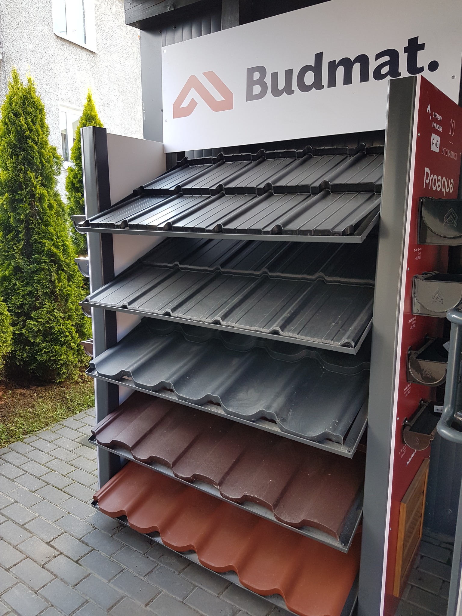 Ekspozycja różnych kolorów i wzorów blachodachówki marki Budmat, prezentowana na zewnątrz budynku, obok iglaków.