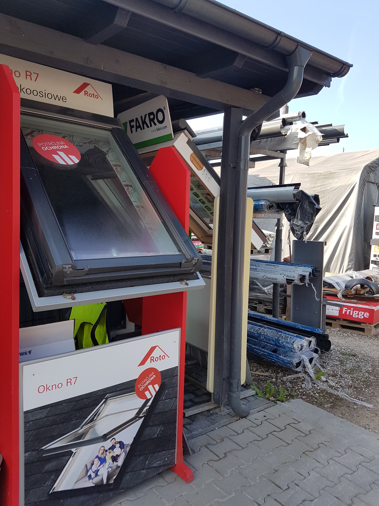 Ekspozycja okien dachowych marek Roto i Fakro z widocznymi rynnami i elementami obróbek blacharskich w tle, prezentowana na zewnątrz budynku w dzień.