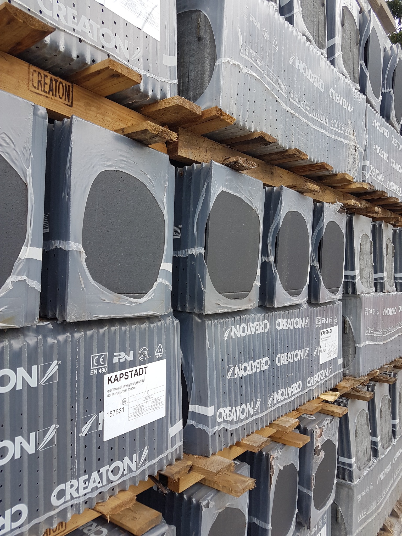 Palety z dachówką ceramiczną CREATON KAPSTADT w grafitowym kolorze, zabezpieczone folią, ułożone jedna na drugiej z drewnianymi przekładkami.