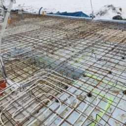 Zbrojenie fundamentu z ułożonymi rurami ogrzewania podłogowego na styropianie, pokryte śniegiem, z widocznymi domami w tle.